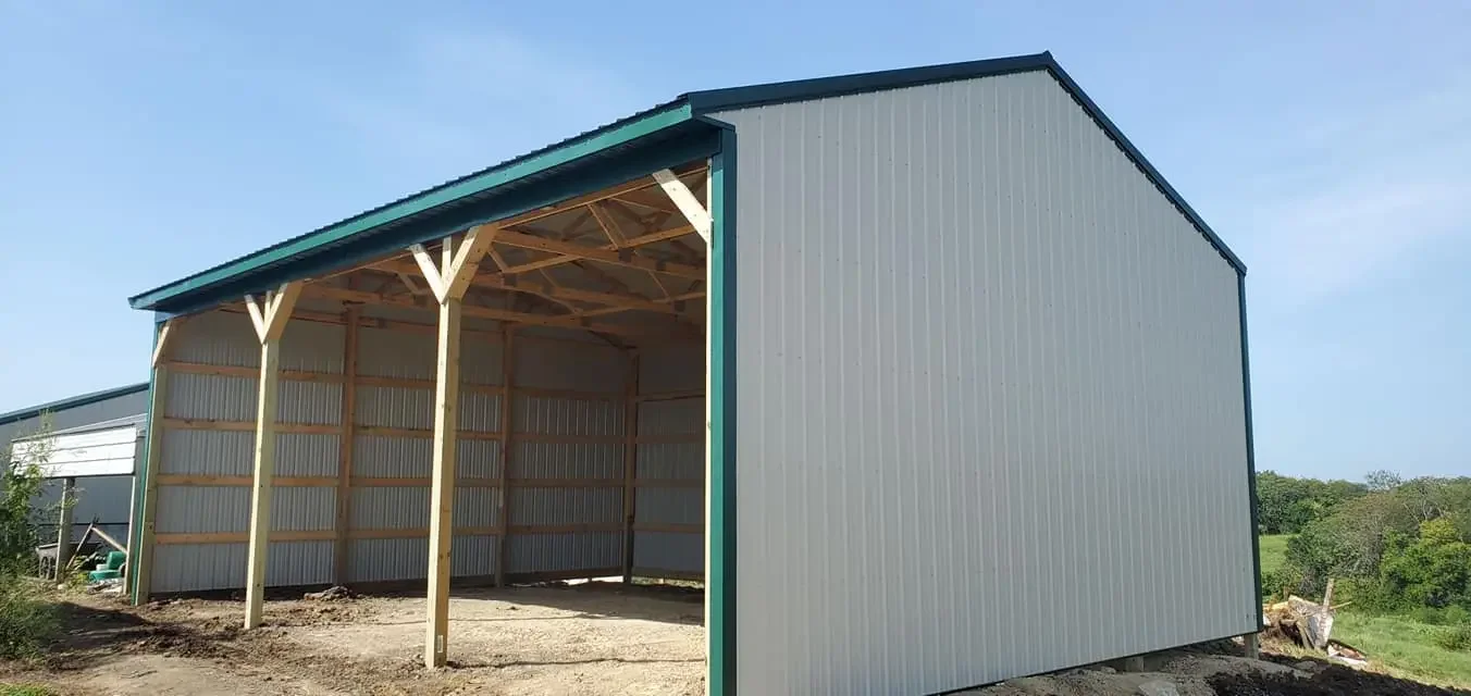 A metal building under construction with an open front and wooden support beams, situated on a dirt patch in a rural area with green trees and grass in the background.