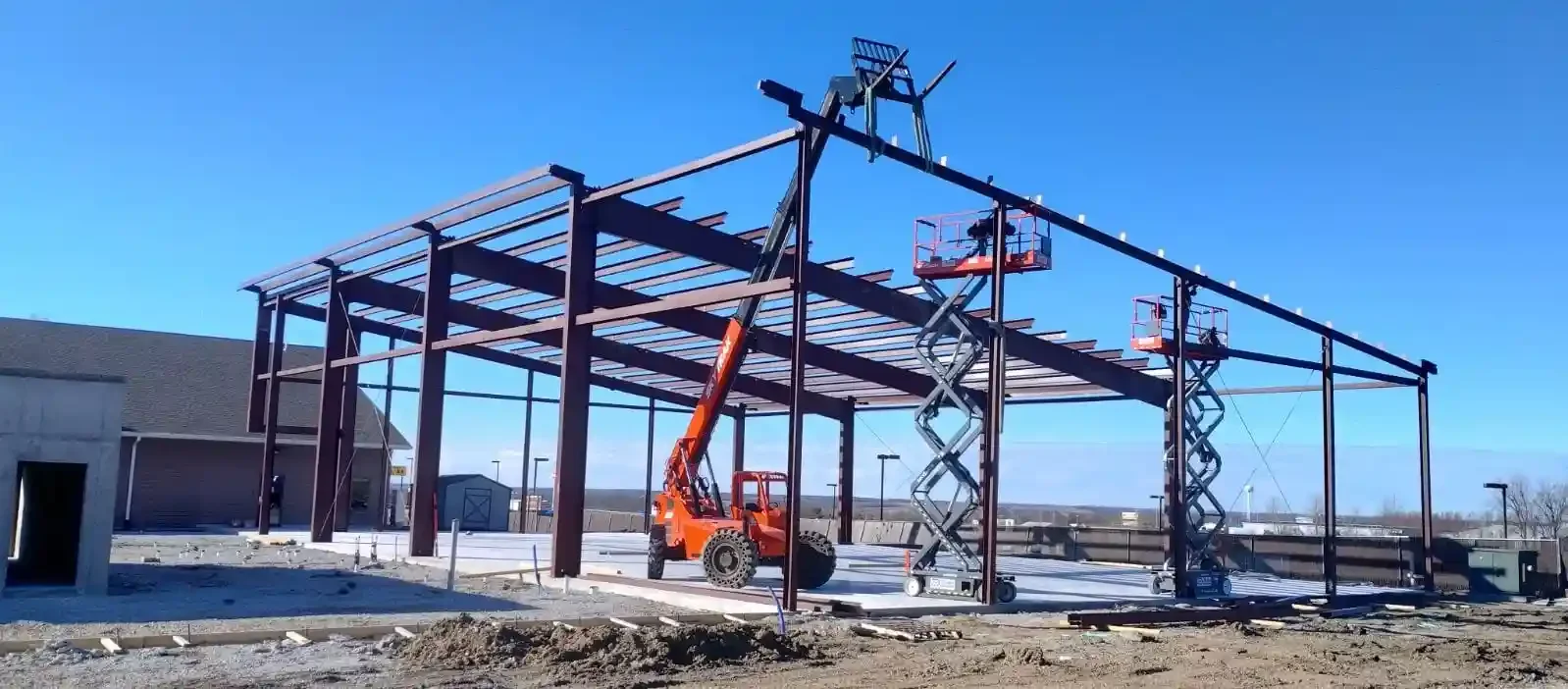 Construction site with a steel frame structure, a lift, and a scissor lift under a clear blue sky.