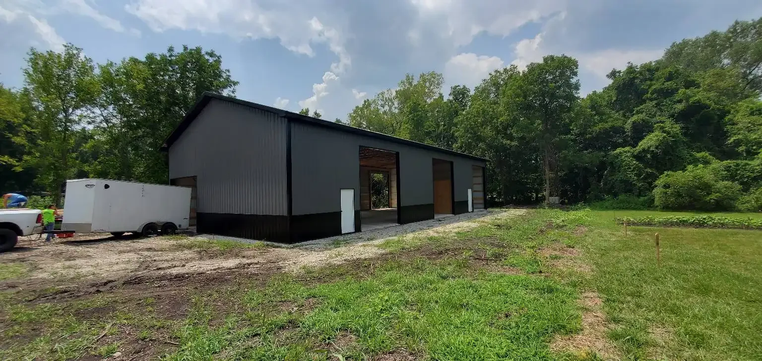 A large grey and black metal building under construction in a rural area with trees in the background, a white trailer parked nearby, and a person in a yellow safety vest.