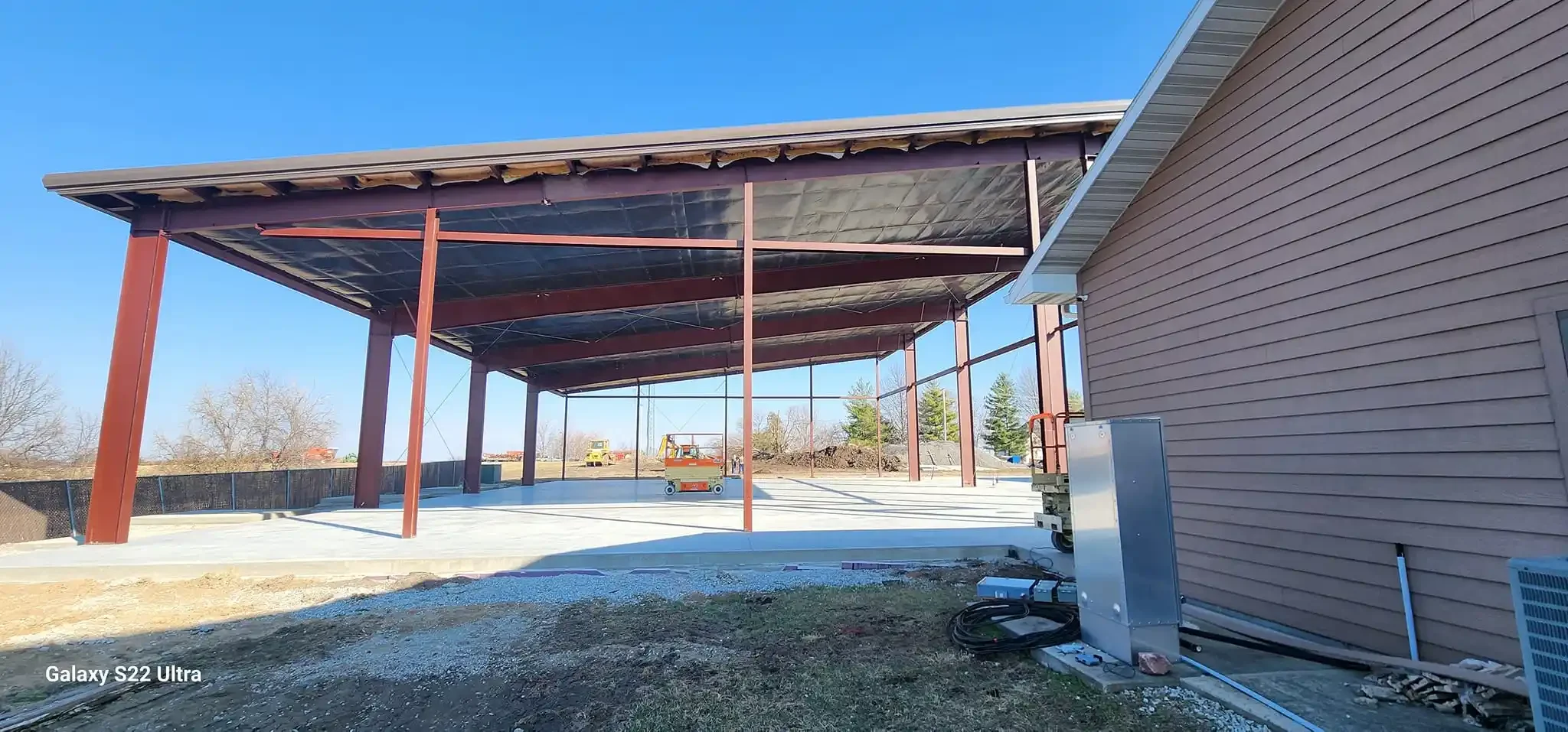 Construction site with a partially built structure featuring steel beams and a concrete floor, adjacent to a house with siding. Equipment and tools are scattered around.