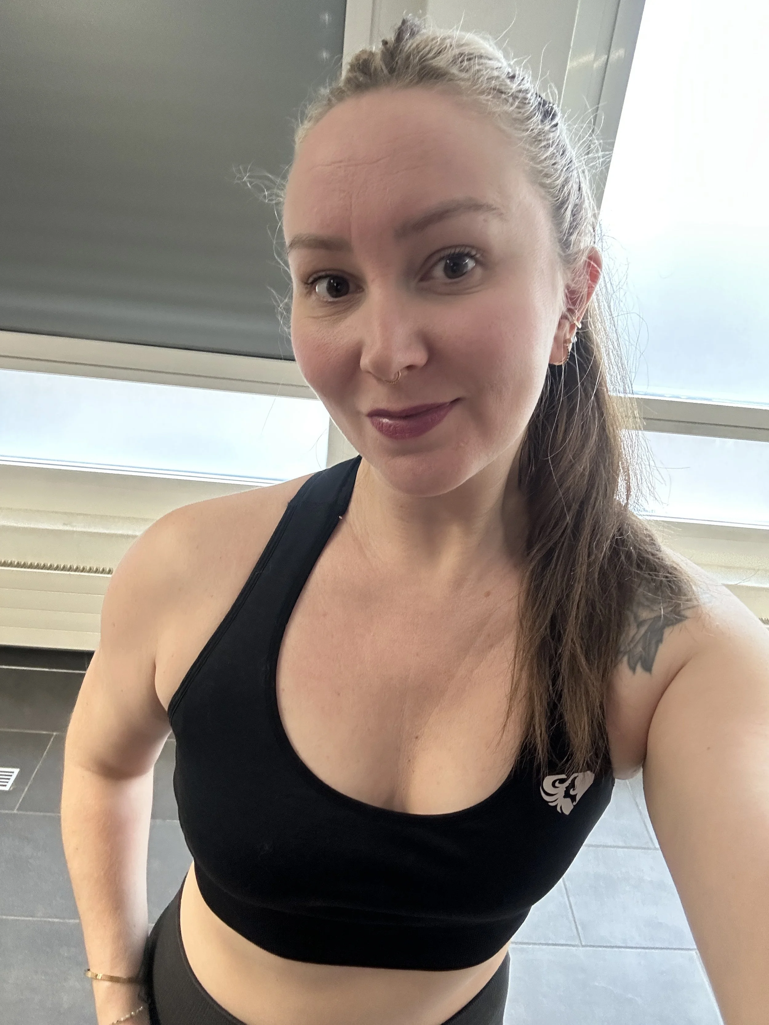 Une jeune femme souriante portant un débardeur noir avec une petite broderie blanche, dans une salle de sport. Elle a une mèche de cheveux bouclée et une peau claire.