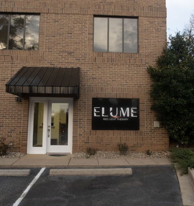 Brick building with sign for ELUME Red Light Therapy, white front door, black awning, parking lot in front.