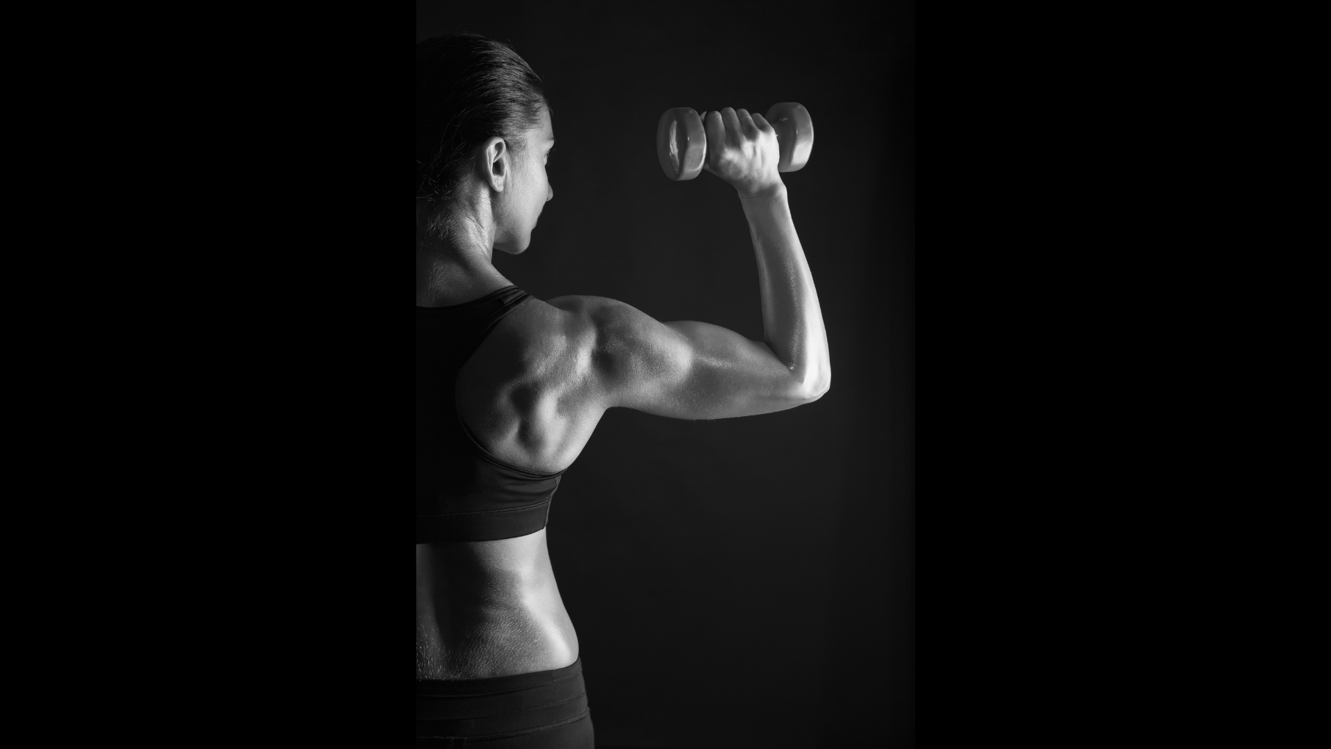 A black and white photo of a woman lifting a dumbbell with her right arm, showing her muscular shoulder and arm, against a dark background.