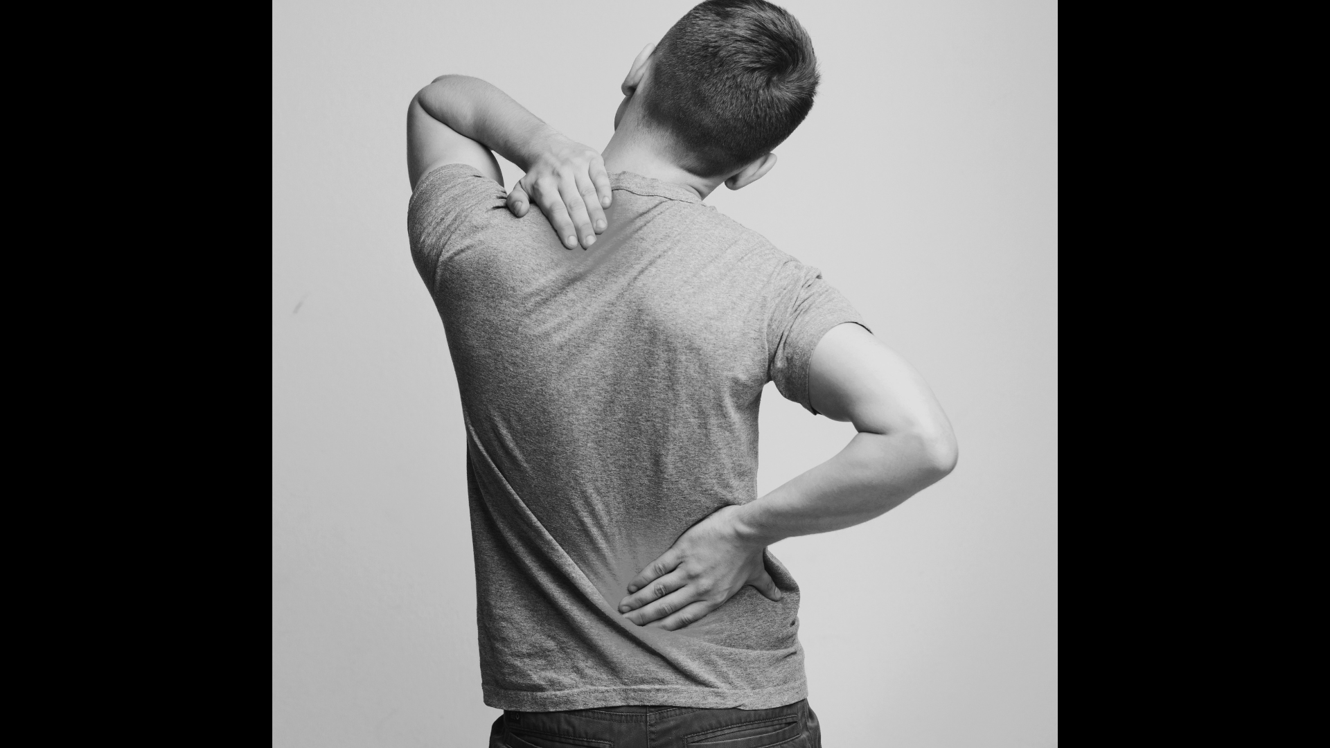 A person with short hair, wearing a gray t-shirt, is seen from the back, touching their shoulder with one hand and placing the other hand on their lower back, appearing to experience discomfort or pain.