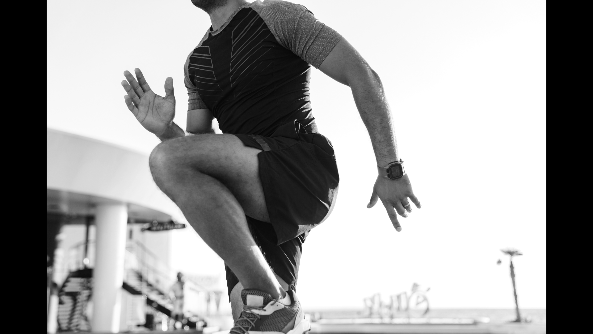 A person in athletic clothing mid-air, performing a jump or exercise outdoors with a clear sky, some structures, and a palm tree in the background.