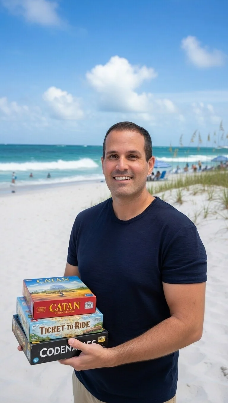 A smiling man in a navy T-shirt holding four board games on a beach with sand, ocean, and blue sky with clouds.
