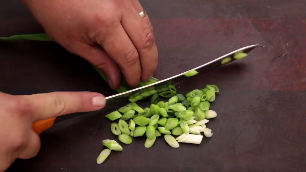 Person slicing green onions on a dark cutting board with a chef's knife.