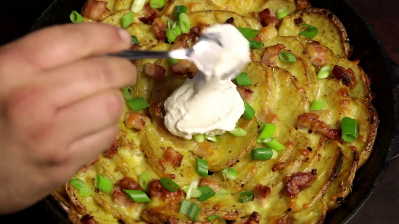 A person is adding sour cream to a dish of sliced potatoes topped with bacon and green onions in a cast-iron skillet.