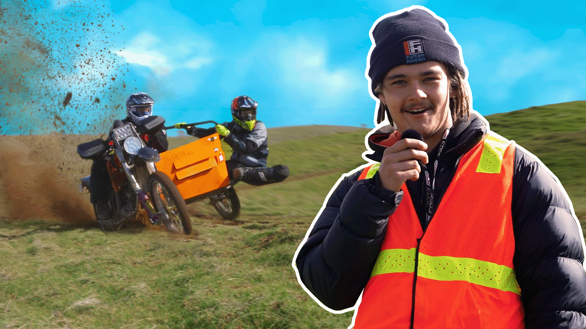 A man in a bright orange safety vest and a dark beanie holds a microphone, smiling, outdoors on a grassy hill. In the background, two motorcyclists wearing helmets and gear are taking a sharp turn, with dirt and grass flying up behind them.