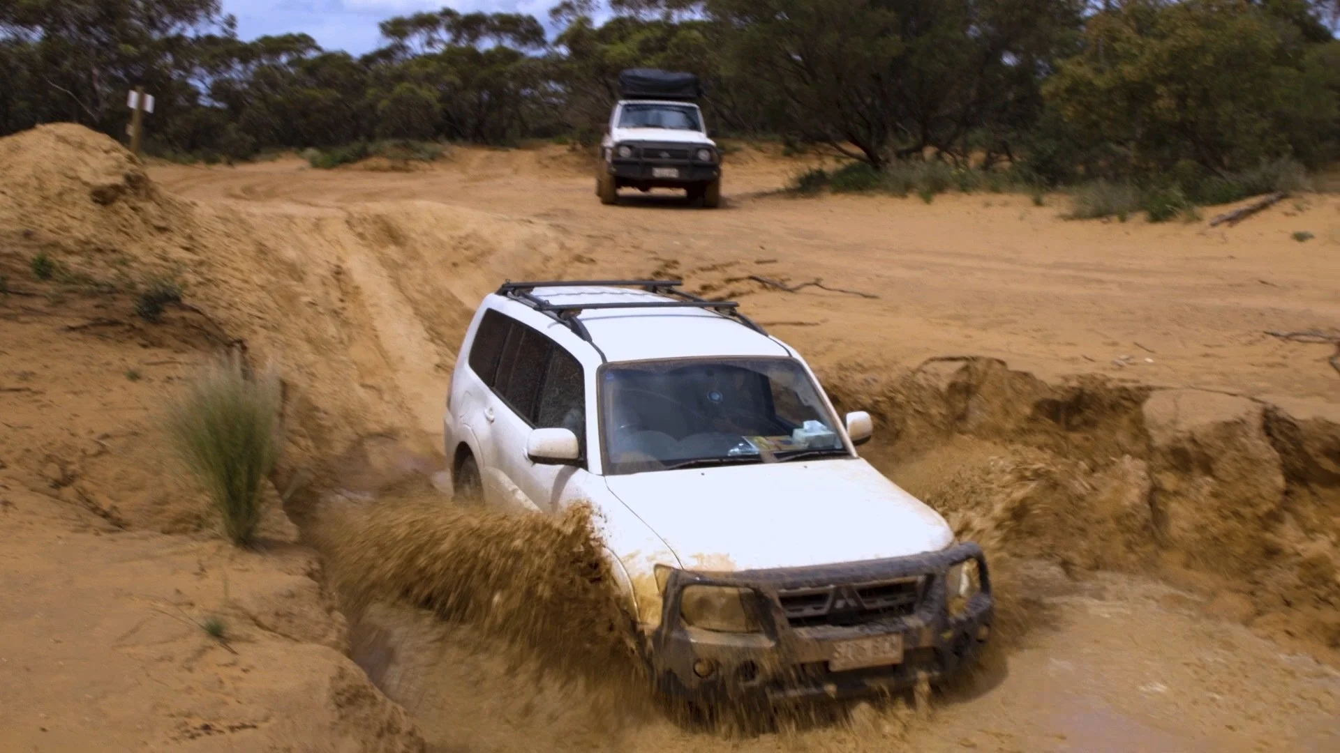 A white SUV driving through a muddy off-road trail, kicking up dirt and mud, with another vehicle parked further along the trail in a rural, wooded area.