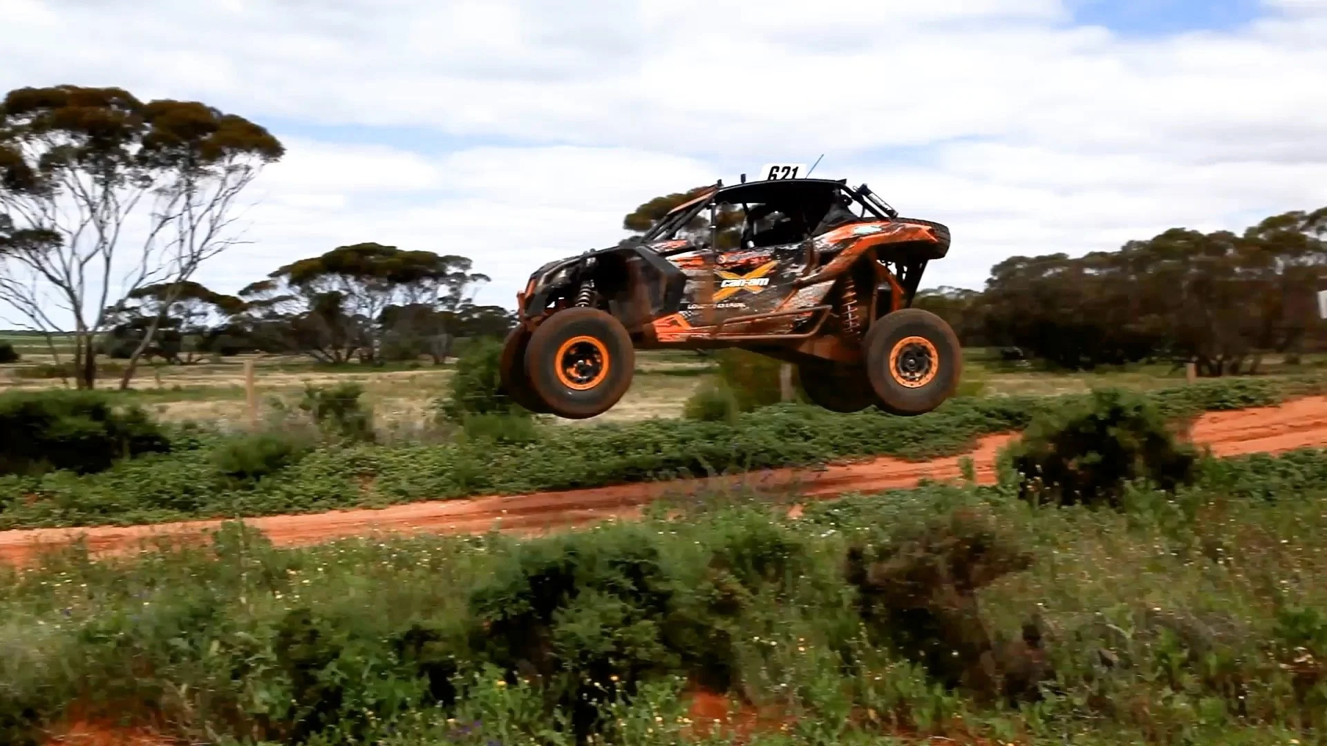 An off-road racing vehicle, orange and black, airborne over dirt and greenery, with a rural landscape and trees in the background.