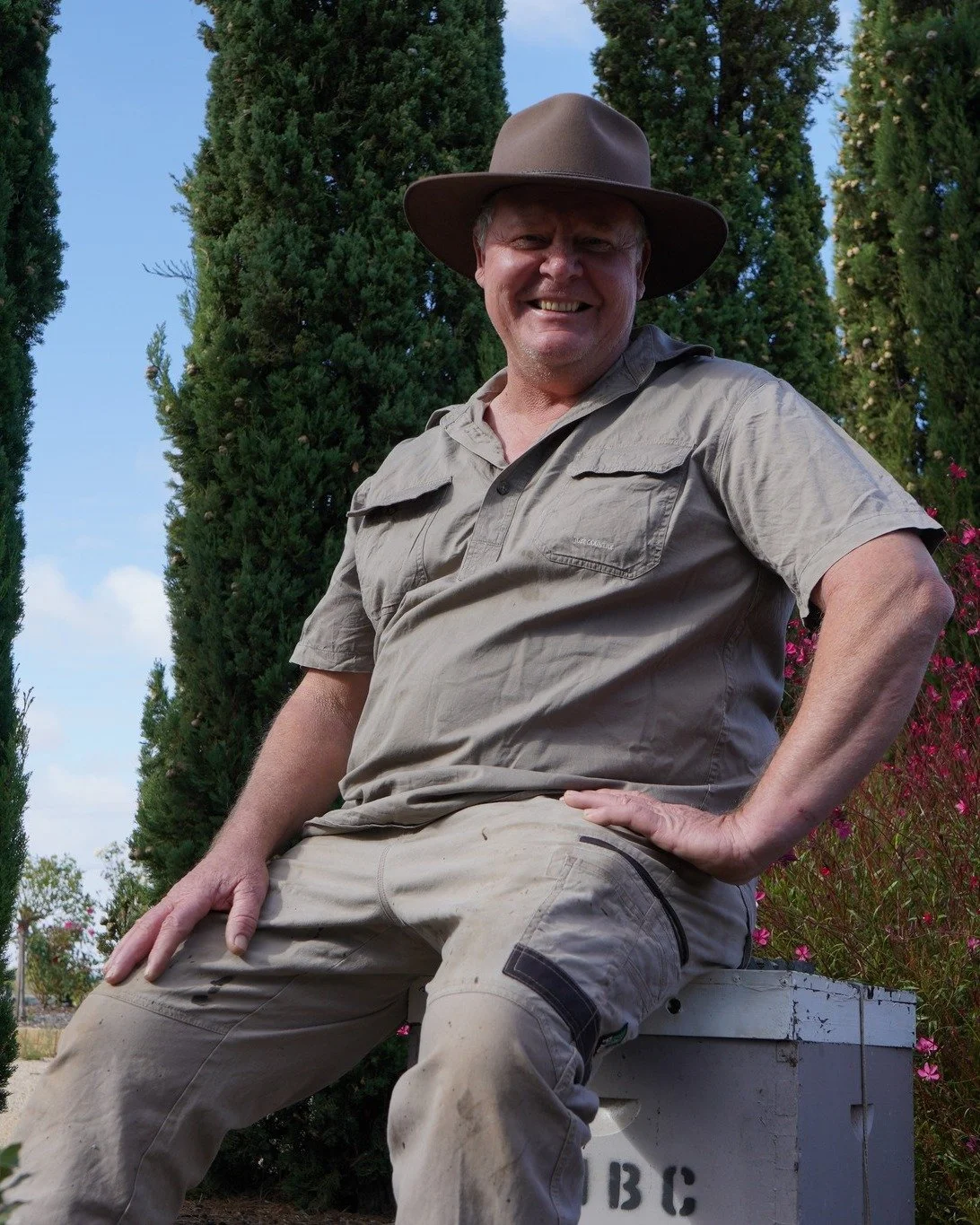A smiling man wearing a wide-brimmed hat and outdoor clothing, sitting outdoors in front of tall green trees and pink flowers.