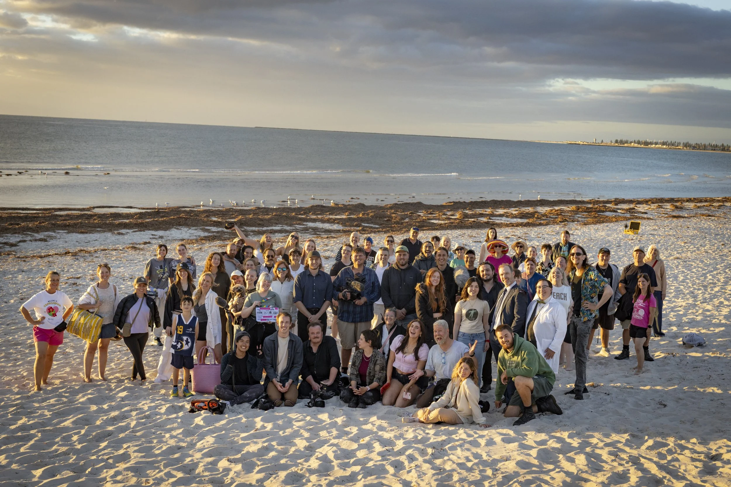 A large group of people on a sandy beach during sunset, with the ocean and a cloudy sky in the background.