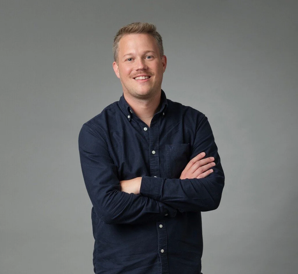 A young man with blonde hair, wearing a dark blue button-up shirt, smiling and crossing his arms, standing against a neutral gray background.