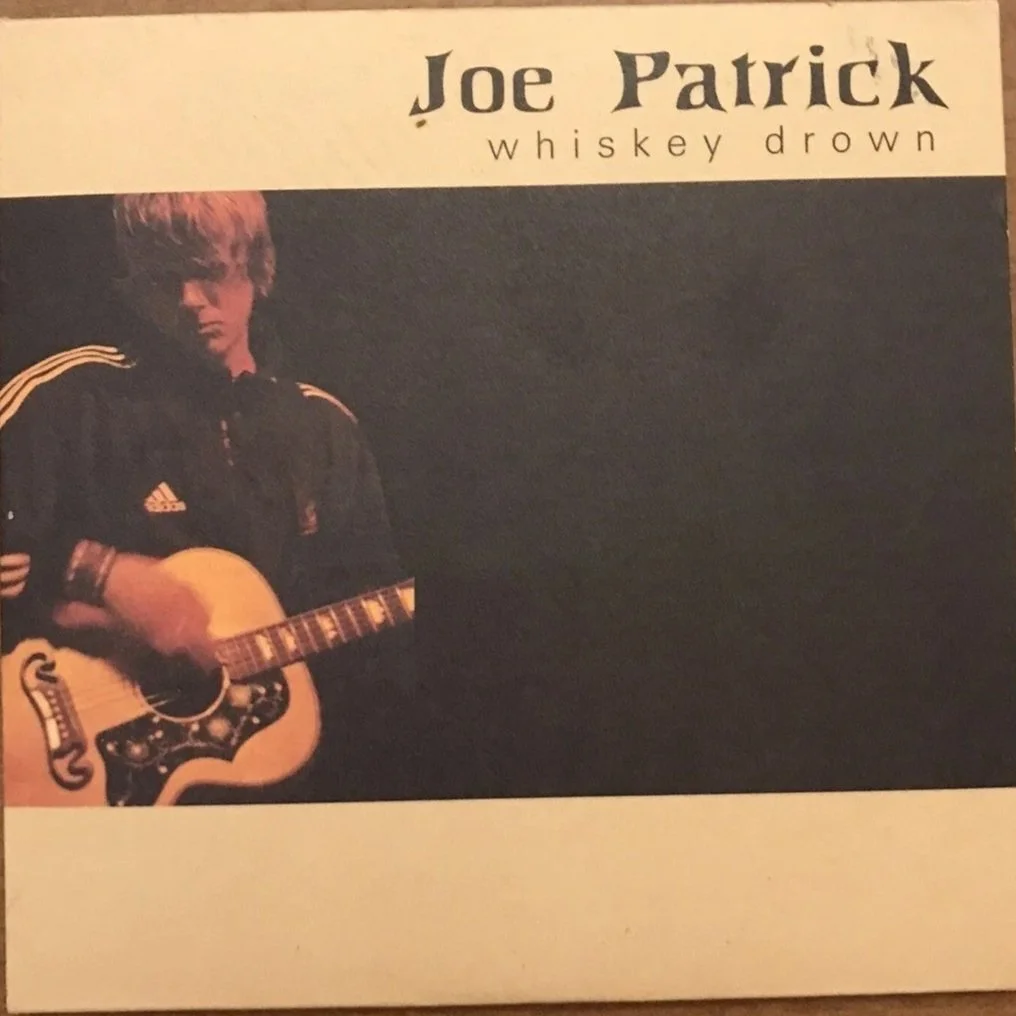 Album cover featuring a young man with blonde hair playing an acoustic guitar, wearing a black jacket with yellow stripes, against a dark background. Text reads 'Joe Patrick Whiskey Drowned'.