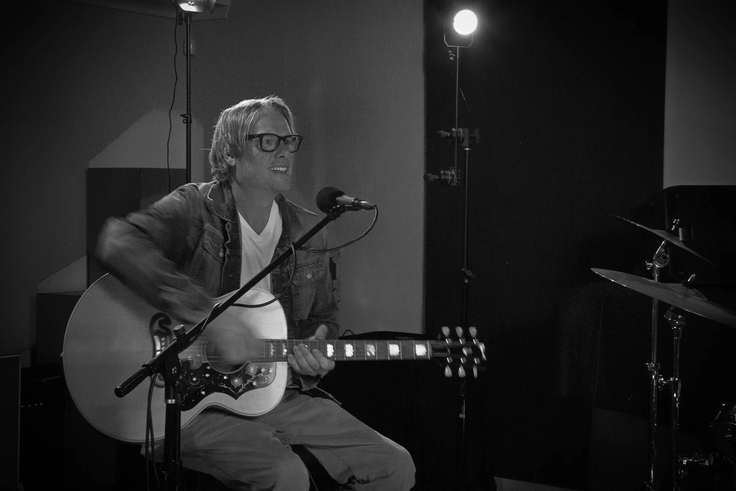 A man with glasses sitting and playing an acoustic guitar in a music studio with a microphone in front of him, black and white photo.