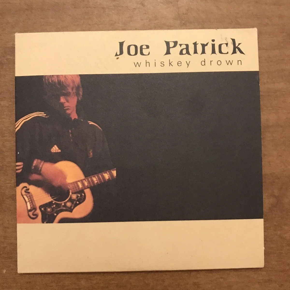 Album cover for Joe Patrick's "Whiskey Drown" featuring a young man with shaggy hair playing an acoustic guitar on a dark background.