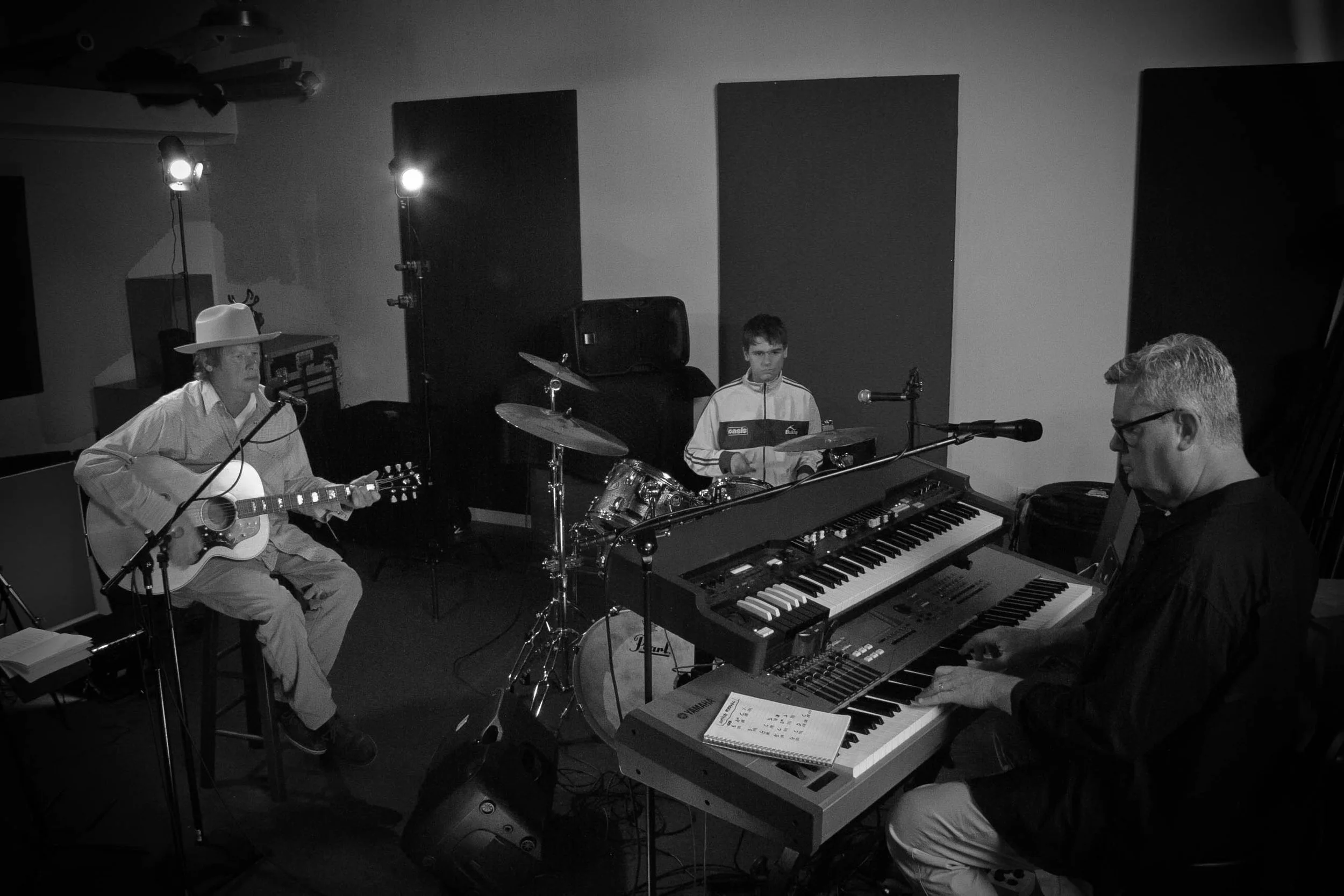 Three musicians in a recording studio: one playing an acoustic guitar and singing, one on drums, and another on an electronic keyboard.