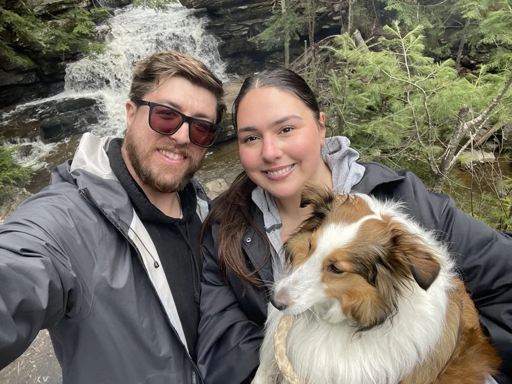 A happy couple with their dog taking a selfie outdoors near a waterfall surrounded by trees.