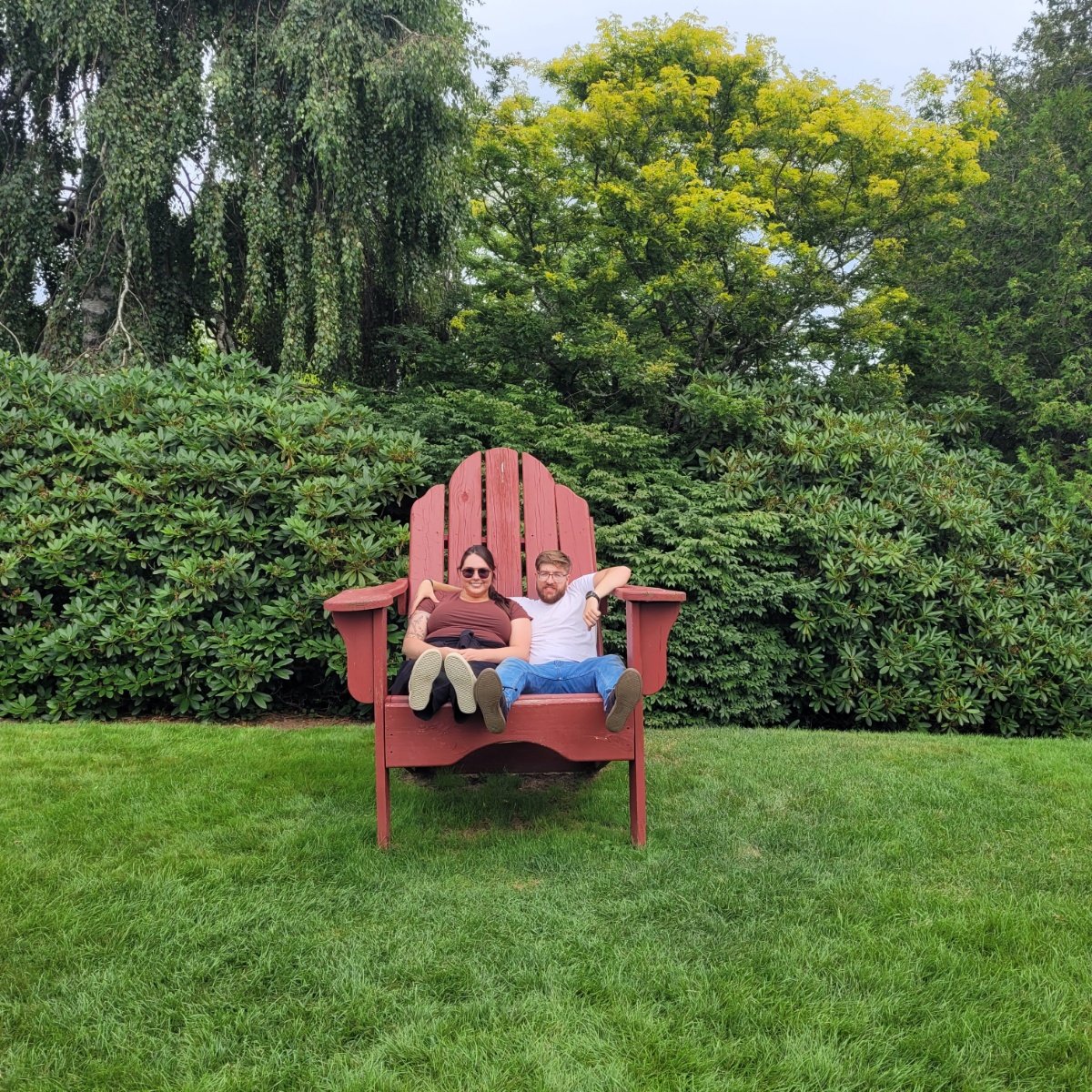 A couple sitting on a large red Adirondack chair outdoors on grass, with green shrubs and trees in the background.