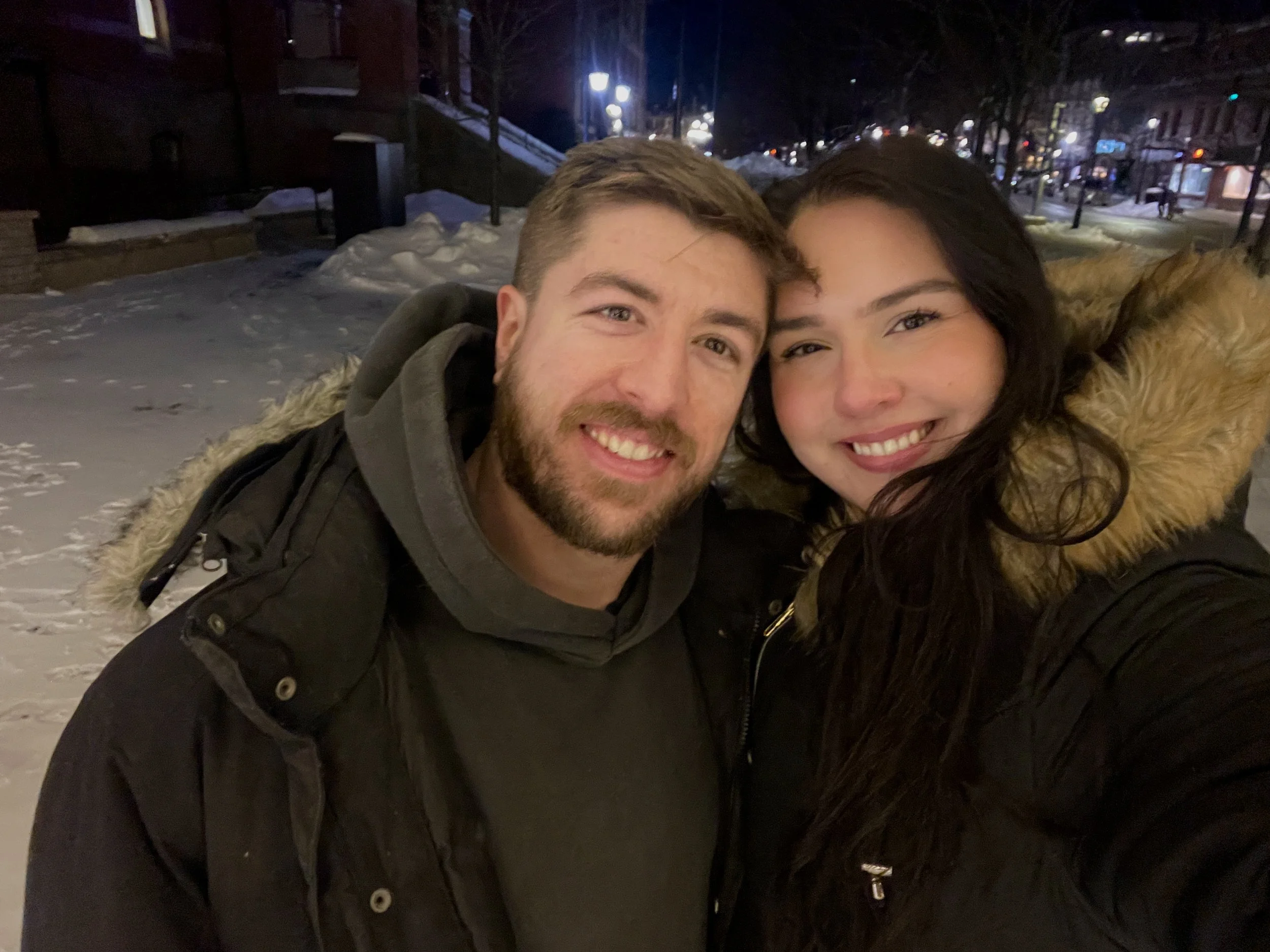 A smiling man and woman taking a selfie outdoors at night in a snowy town, both wearing warm winter coats.