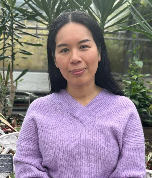 A woman with long black hair smiling, wearing a light purple sweater, positioned outdoors with greenery and plants in the background.