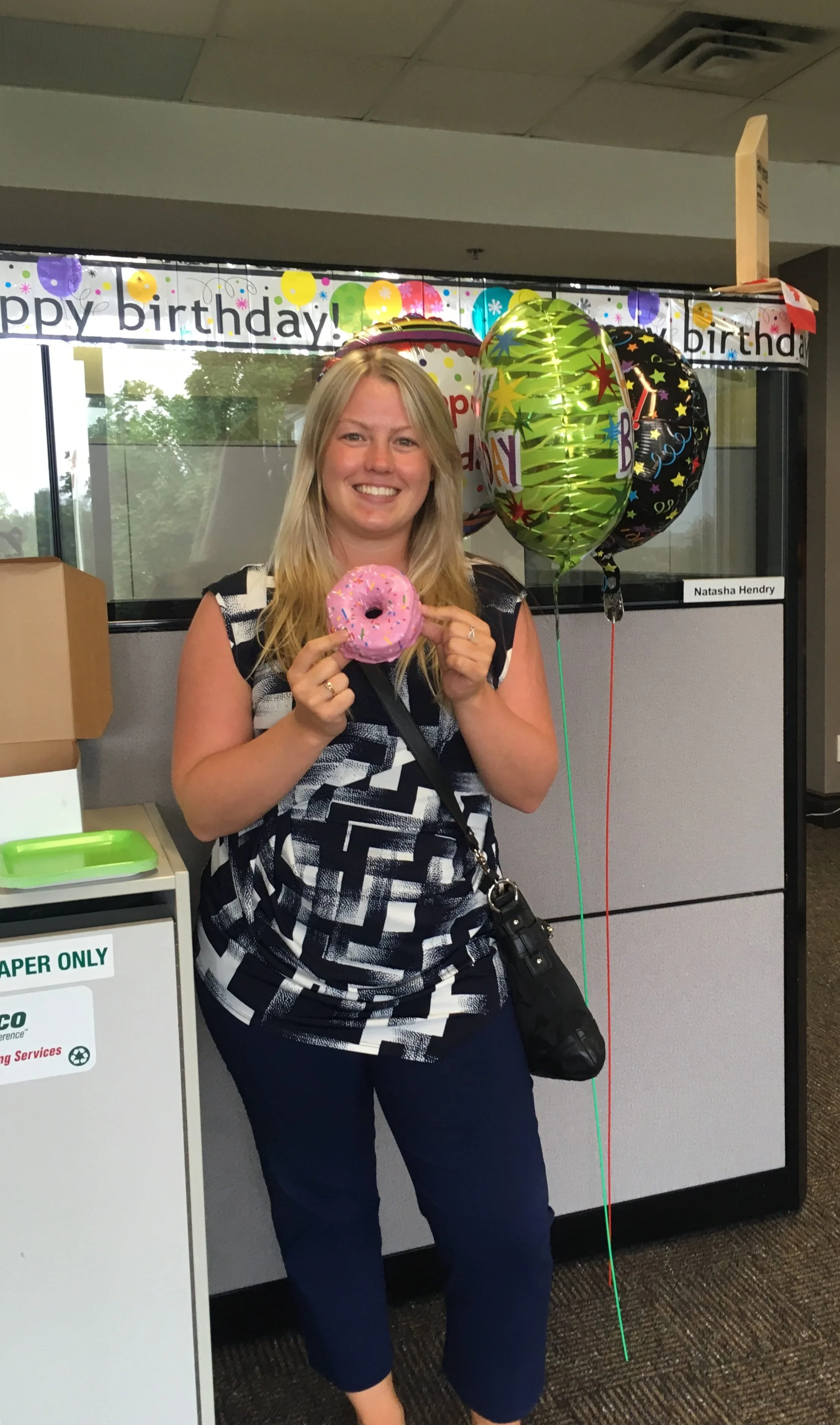 A woman smiling and holding a pink frosted donut with sprinkles, standing in an indoor setting with birthday decorations, including balloons and a banner that says 'happy birthday' in the background.