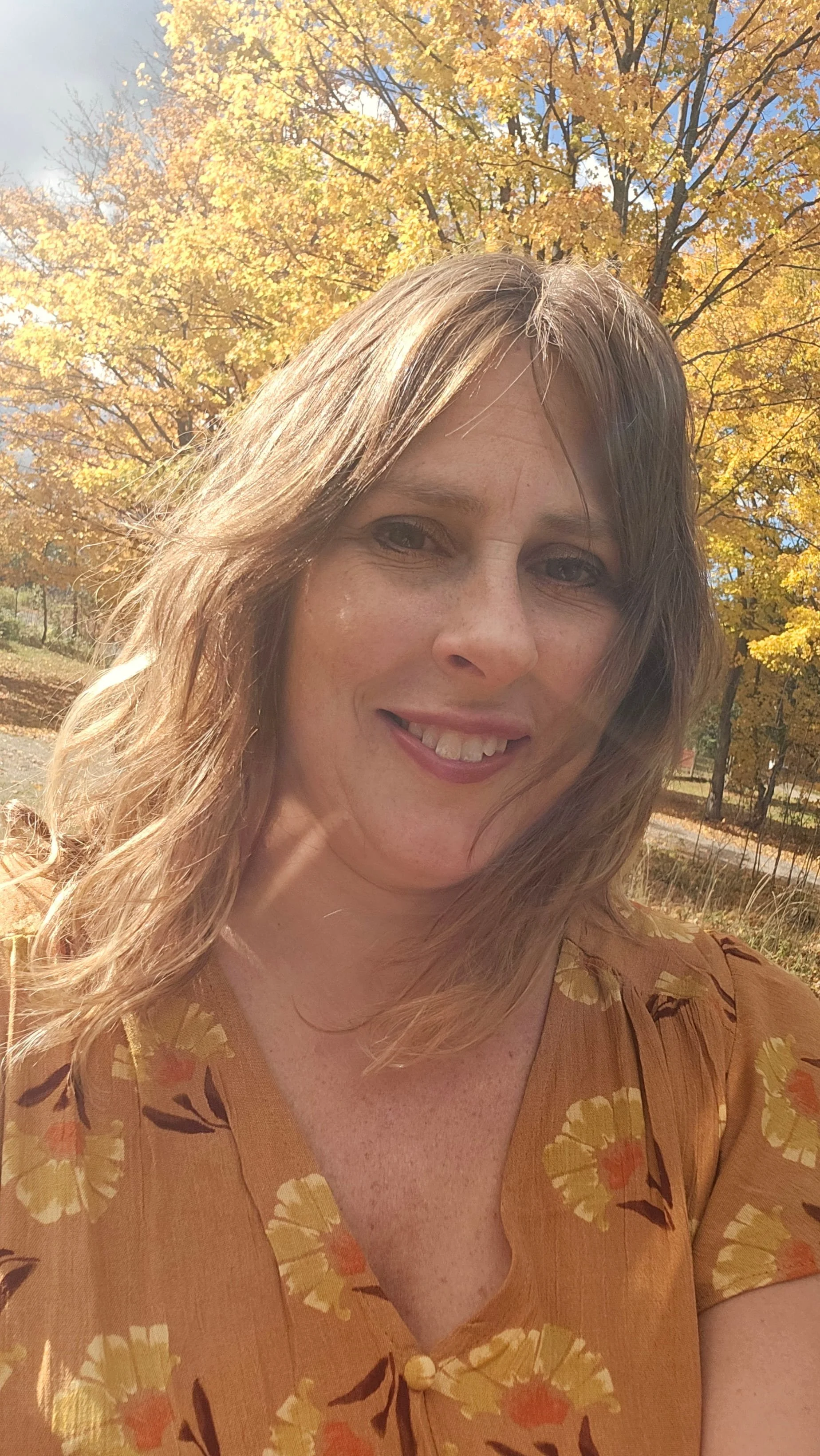 A woman with light brown hair smiling outdoors in front of autumn trees with yellow leaves, wearing a brown floral dress.
