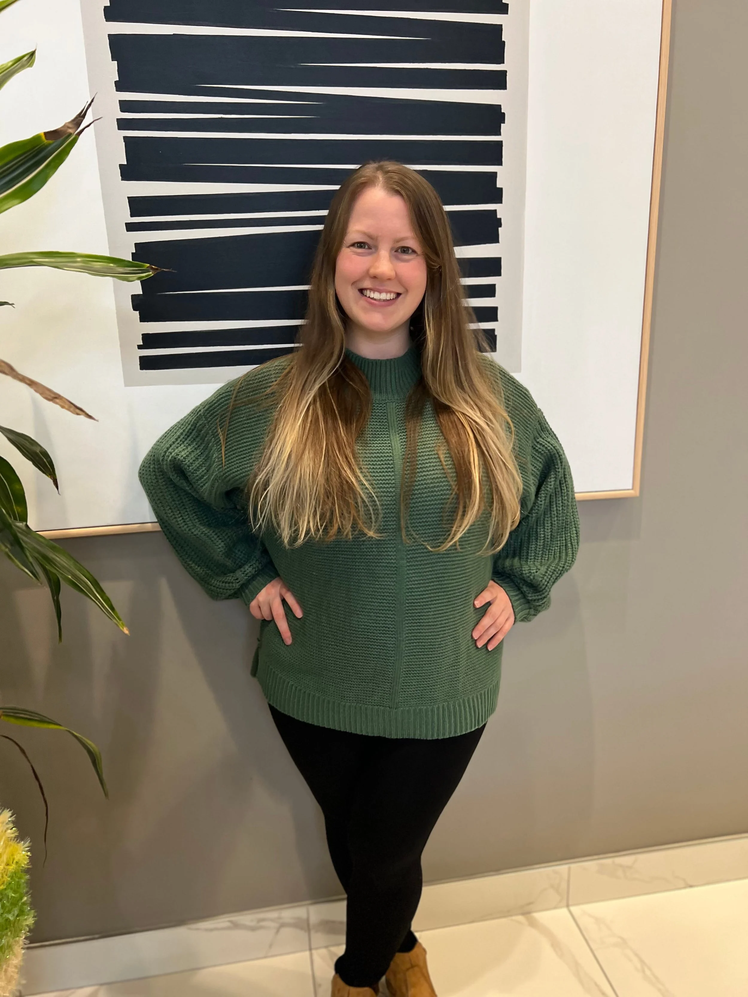 A woman with long, wavy, light brown hair standing indoors in front of a modern abstract black and white artwork on the wall. She is wearing a green knit sweater, black leggings, and tan boots, smiling with hands on hips.