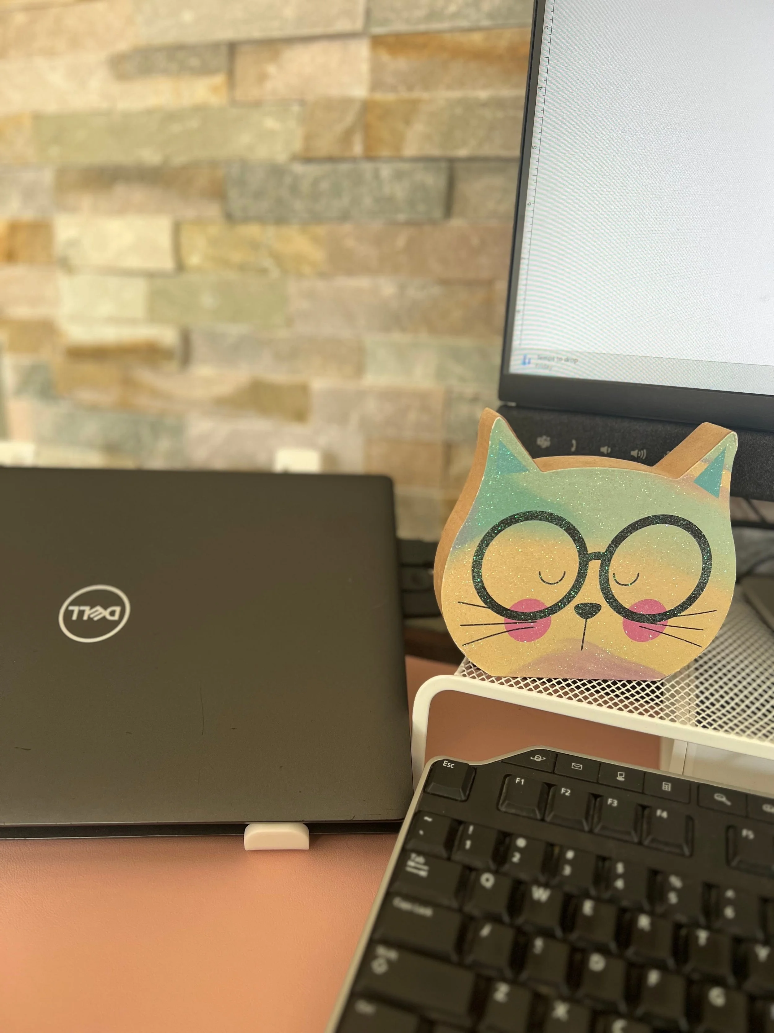 A computer workspace with a Dell laptop, a computer monitor, a keyboard, and a colorful cat-shaped paper decoration on a white wire mesh desk organizer. The background features a brick wall.