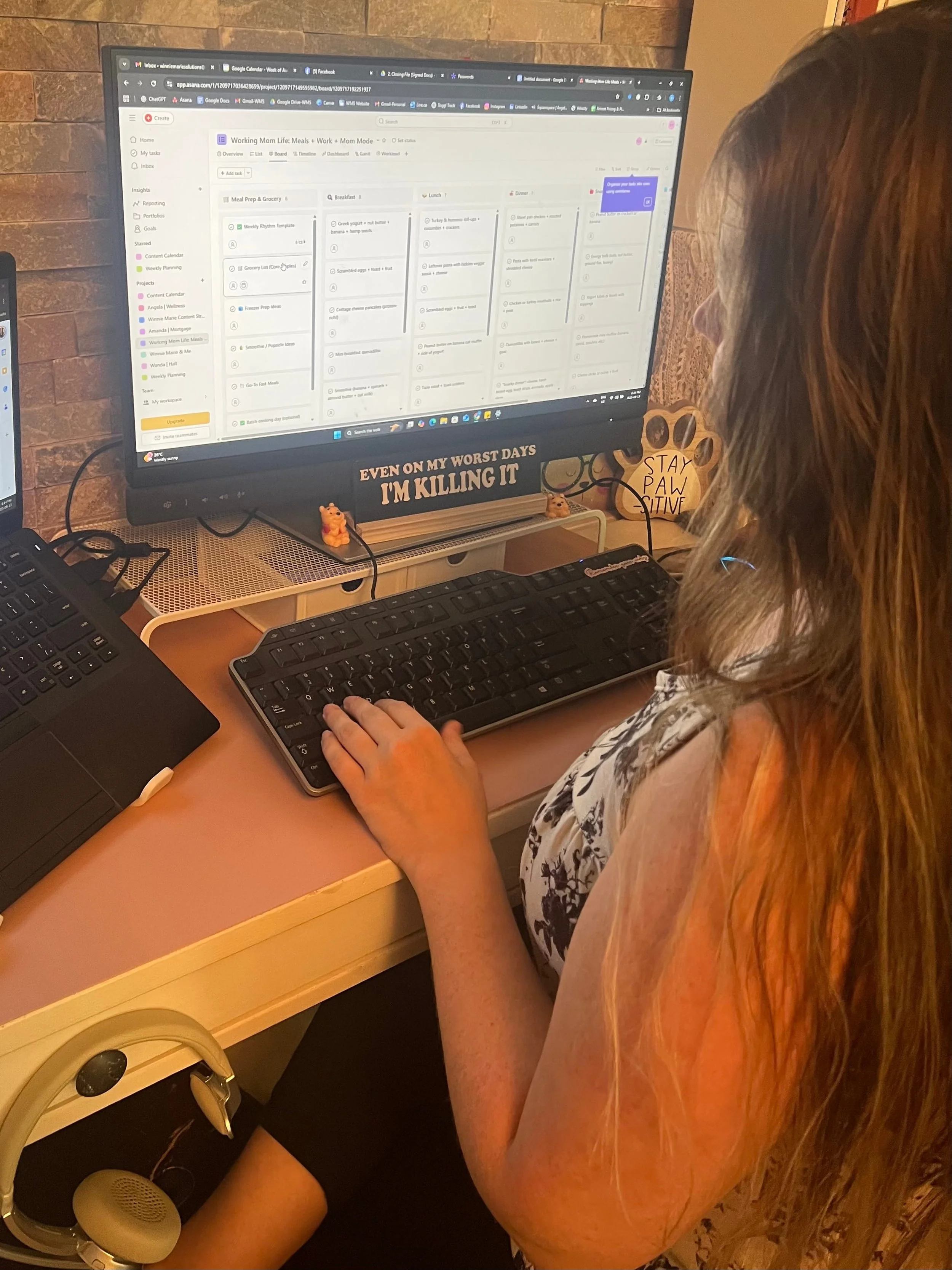 A person with long reddish hair sitting at a desk using a keyboard and looking at a computer monitor displaying a digital planner titled 'Working Mom Life: Meals + Work + Mom Mode.' The desk has a keyboard, a computer mouse, and a small portable speaker. A small sign on the wall behind the desk reads, 'STAY PAW-SITIVE.'