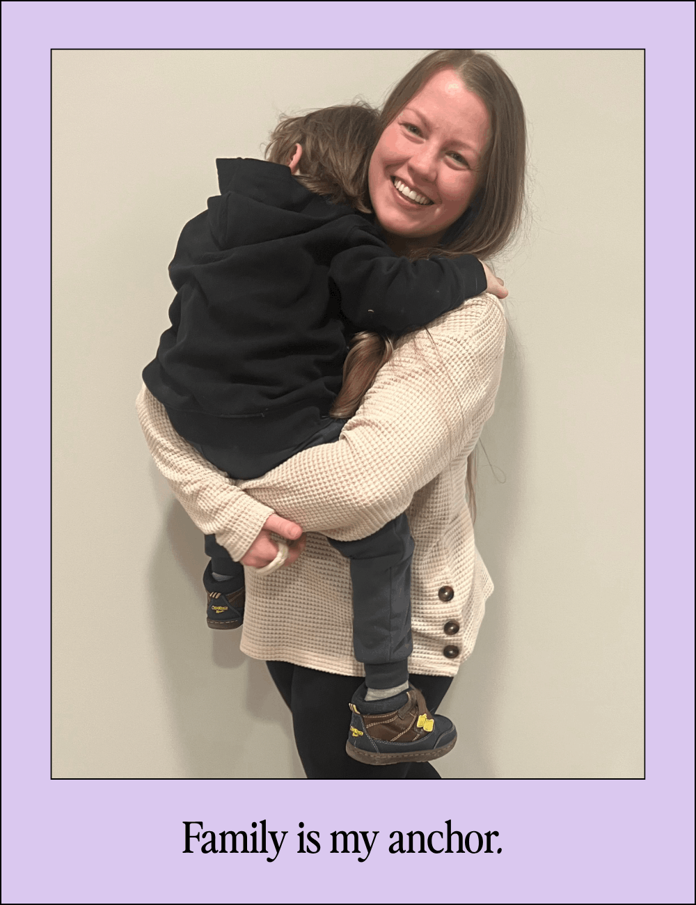 A woman holding a young child in a hug, both smiling against a plain wall.