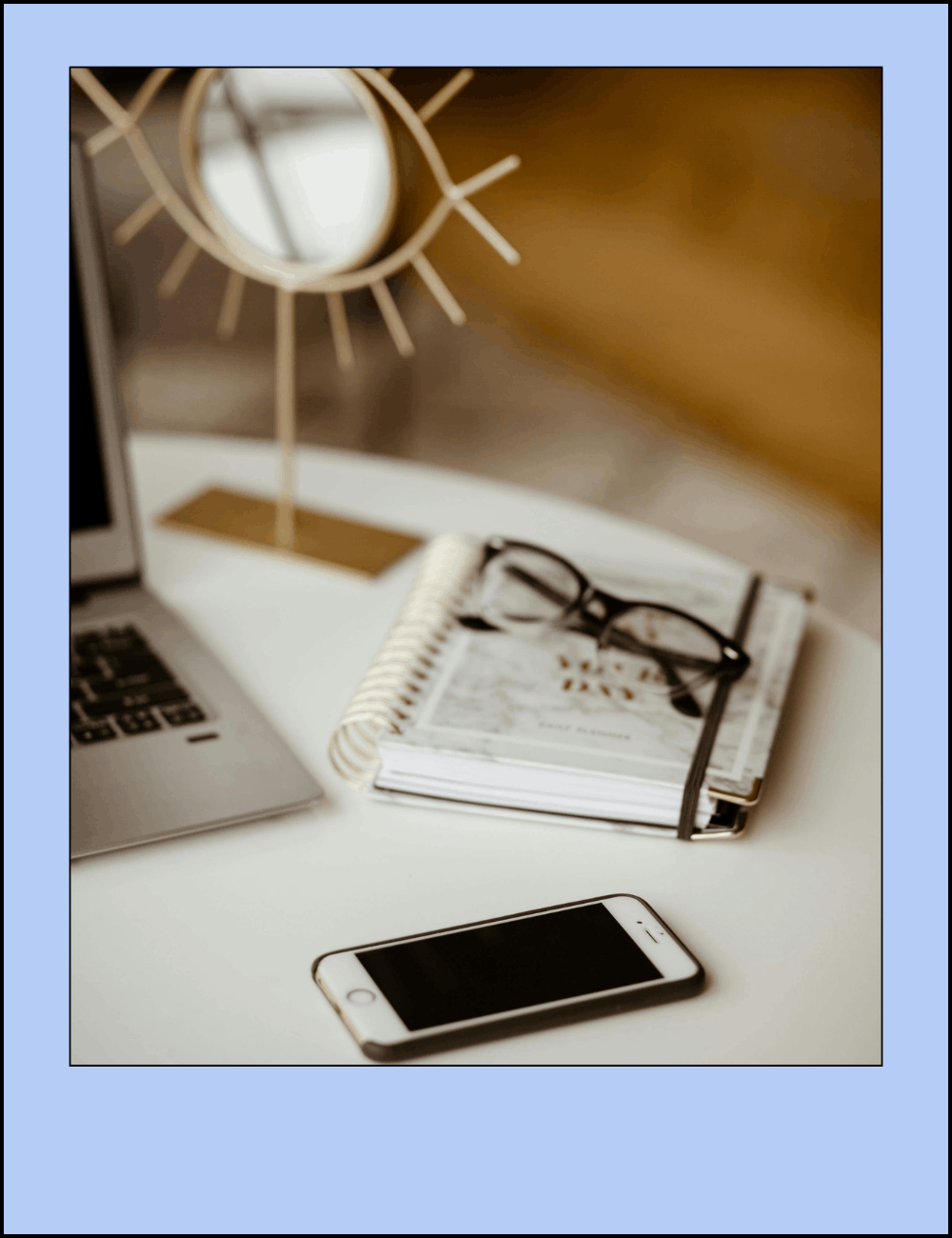 A workspace with a laptop, a smartphone, a spiral notepad with glasses on top, a mirror, and a decorative clock.