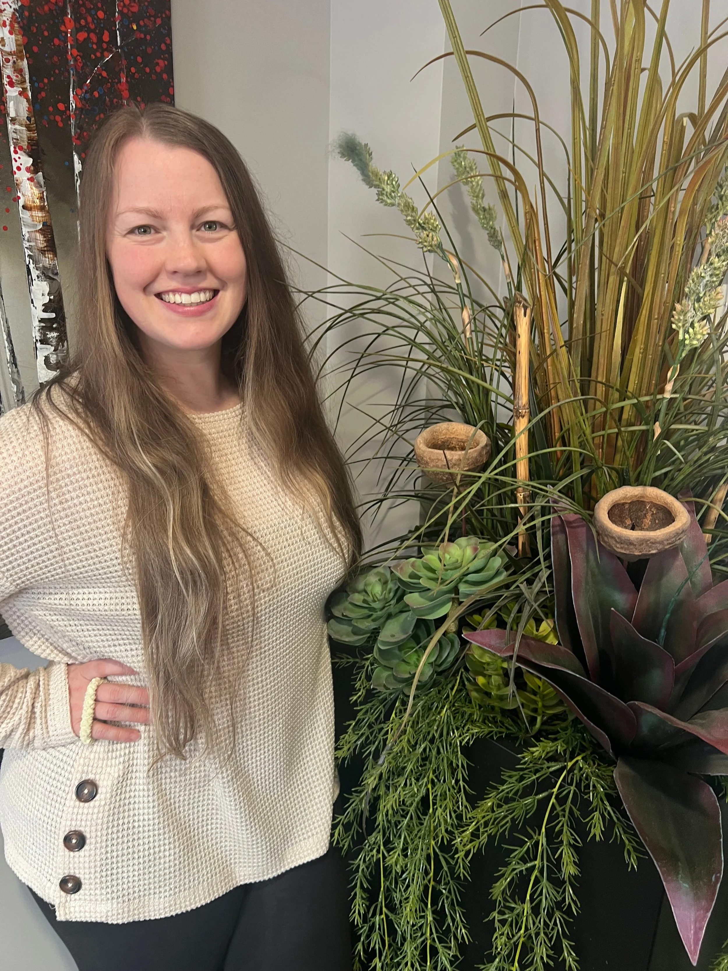 A woman with long brown hair, smiling, wearing a beige sweater, standing next to various indoor plants including a succulent, ferns, and tall grasses, with a colorful abstract art piece on the wall in the background.
