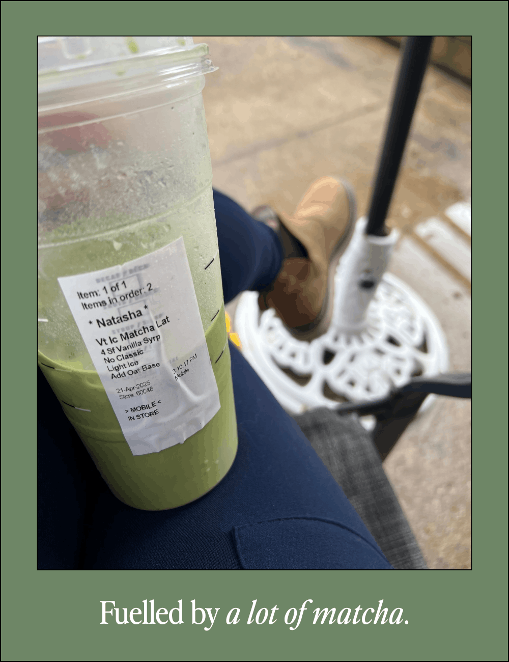 A person sitting outdoors holding a large green matcha latte from Starbucks with a label. The person wears jeans and tan shoes, and there is a parasol or umbrella stand nearby on a paved surface.