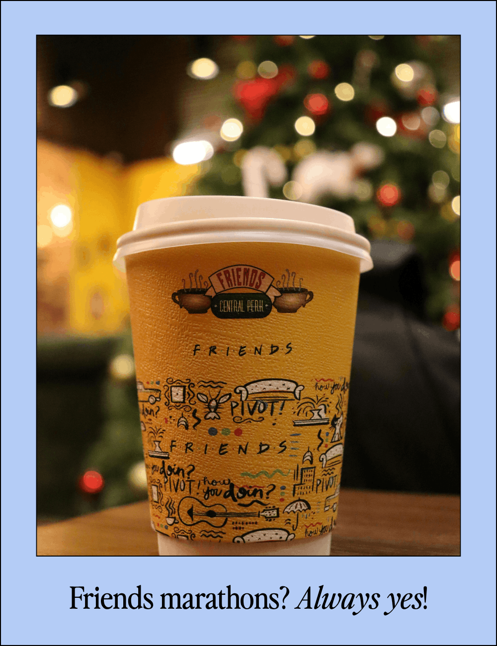 A paper coffee cup with colorful doodles and text, sitting on a wooden table with a blurred Christmas tree with lights in the background.