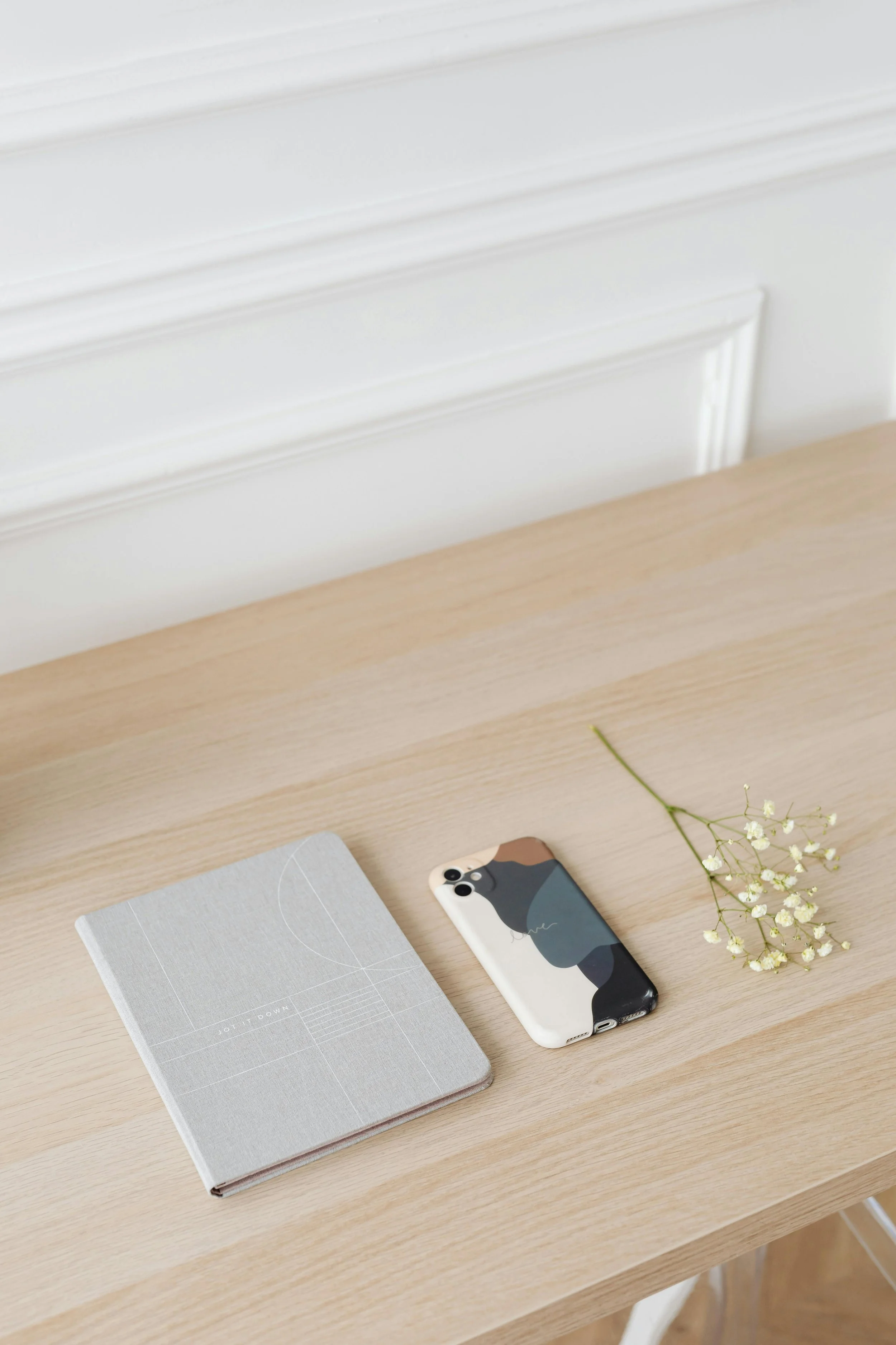 A light wood desk with a gray notebook, a white smartphone with a black, brown, and gray abstract case, and a small branch of white flowers.