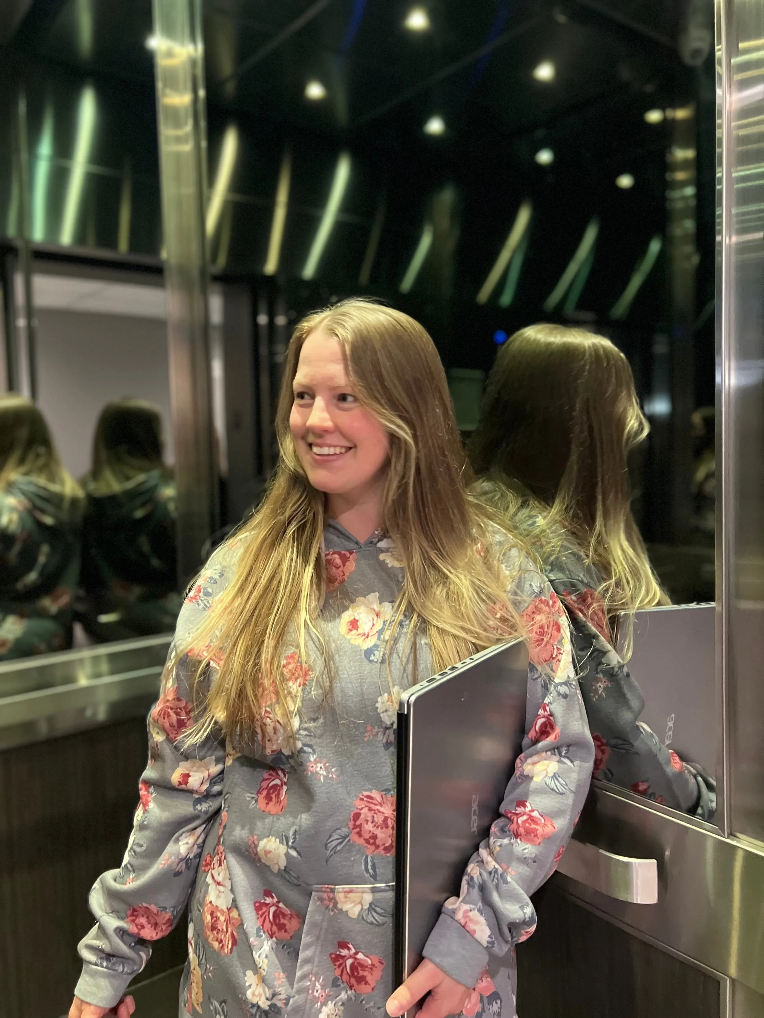 A young woman with long blonde hair smiling inside an elevator with mirrored walls, wearing a gray sweatshirt with a floral pattern, and holding a laptop.