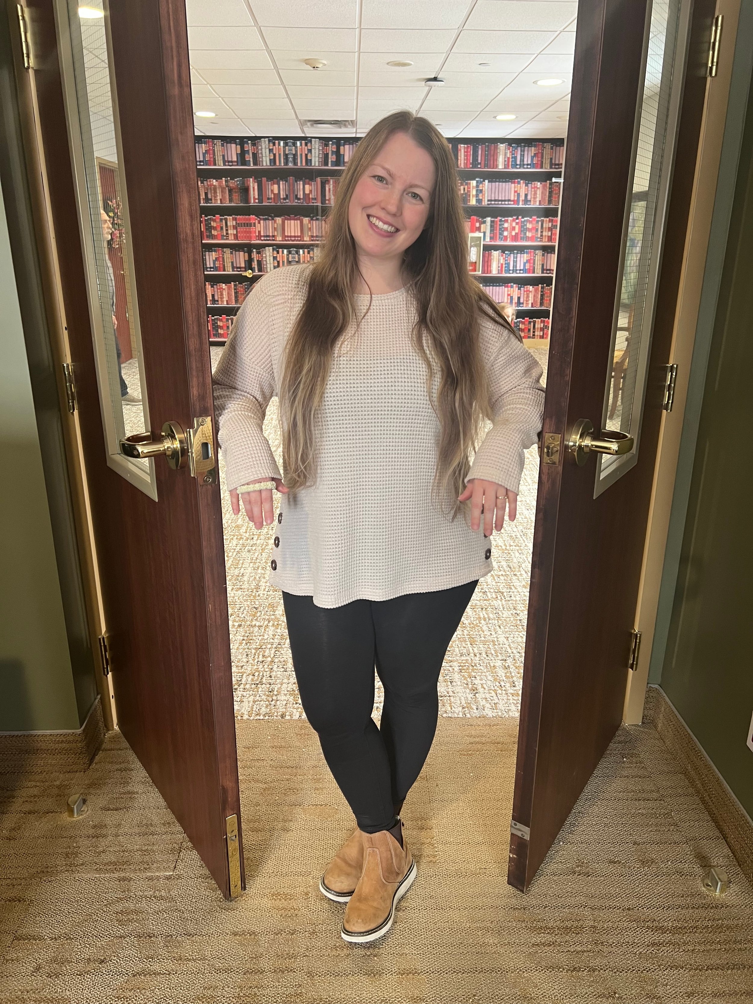 A woman with long brown hair smiling and standing in a doorway with a library background.
