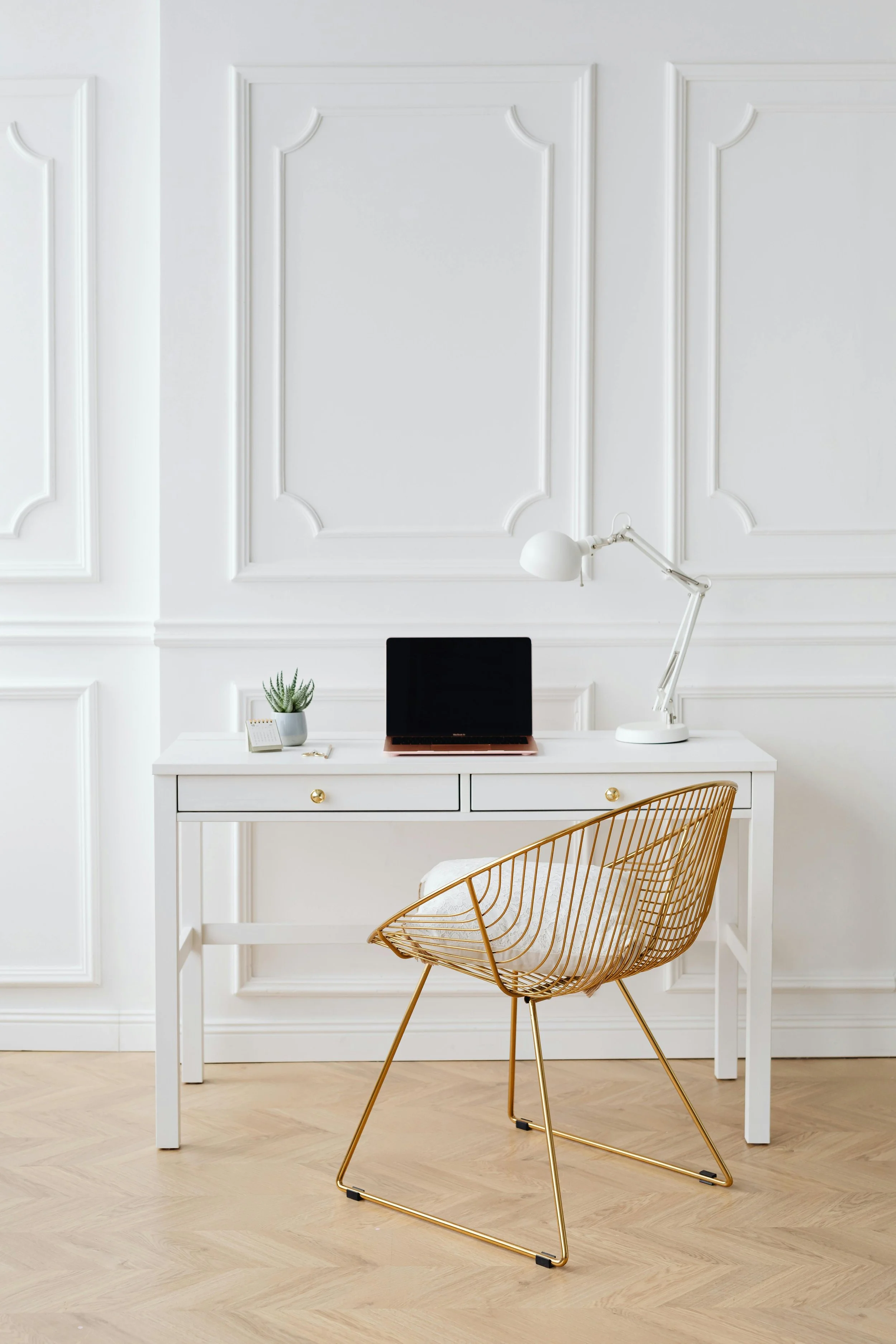 White desk with a computer, a lamp, a small plant, and a notepad, in a bright room with white paneled walls and light wood flooring, gold wire chair in front.