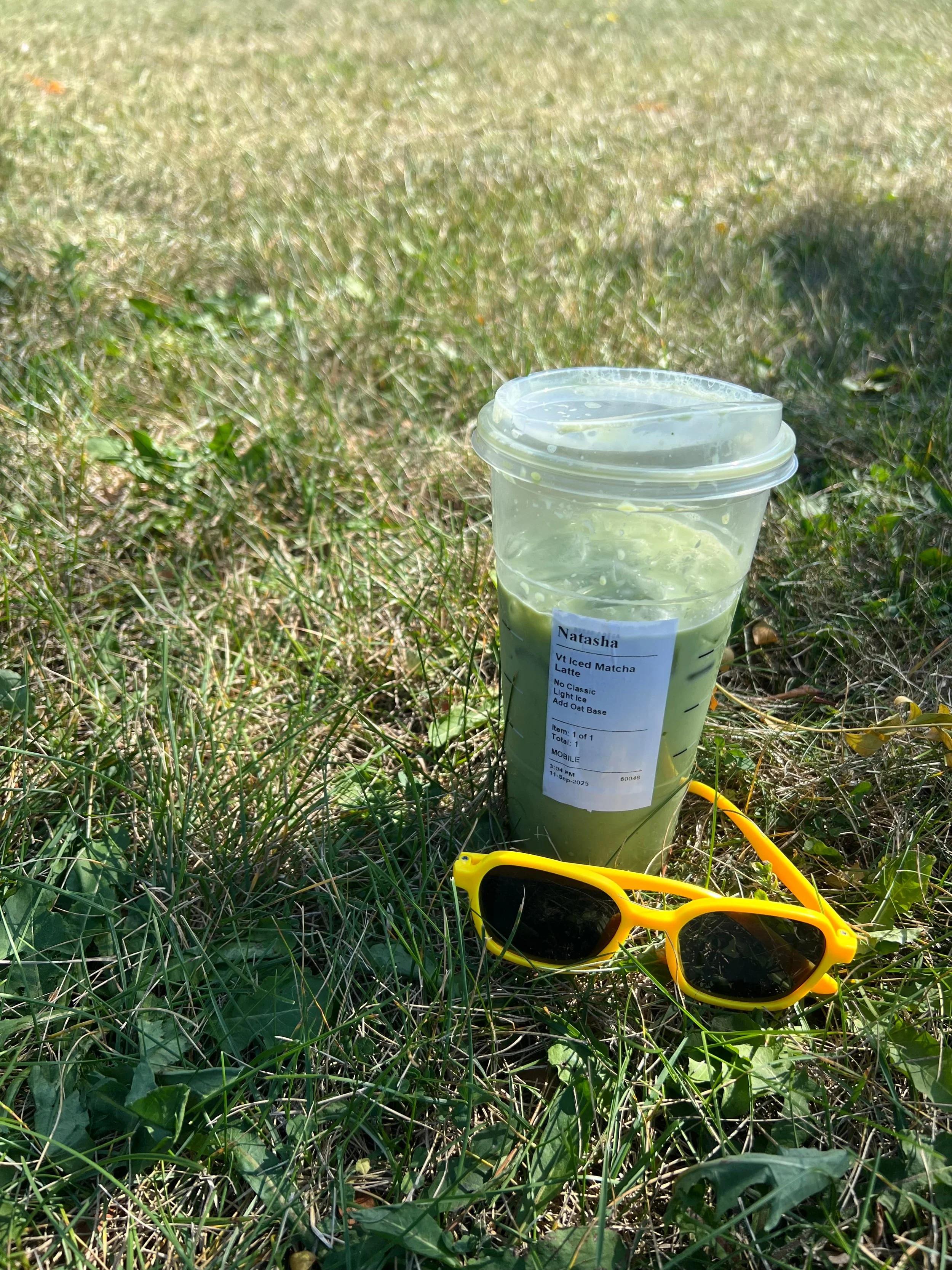A cup of iced matcha latte with a lid, yellow sunglasses, and a hand-held refractometer on grassy ground.