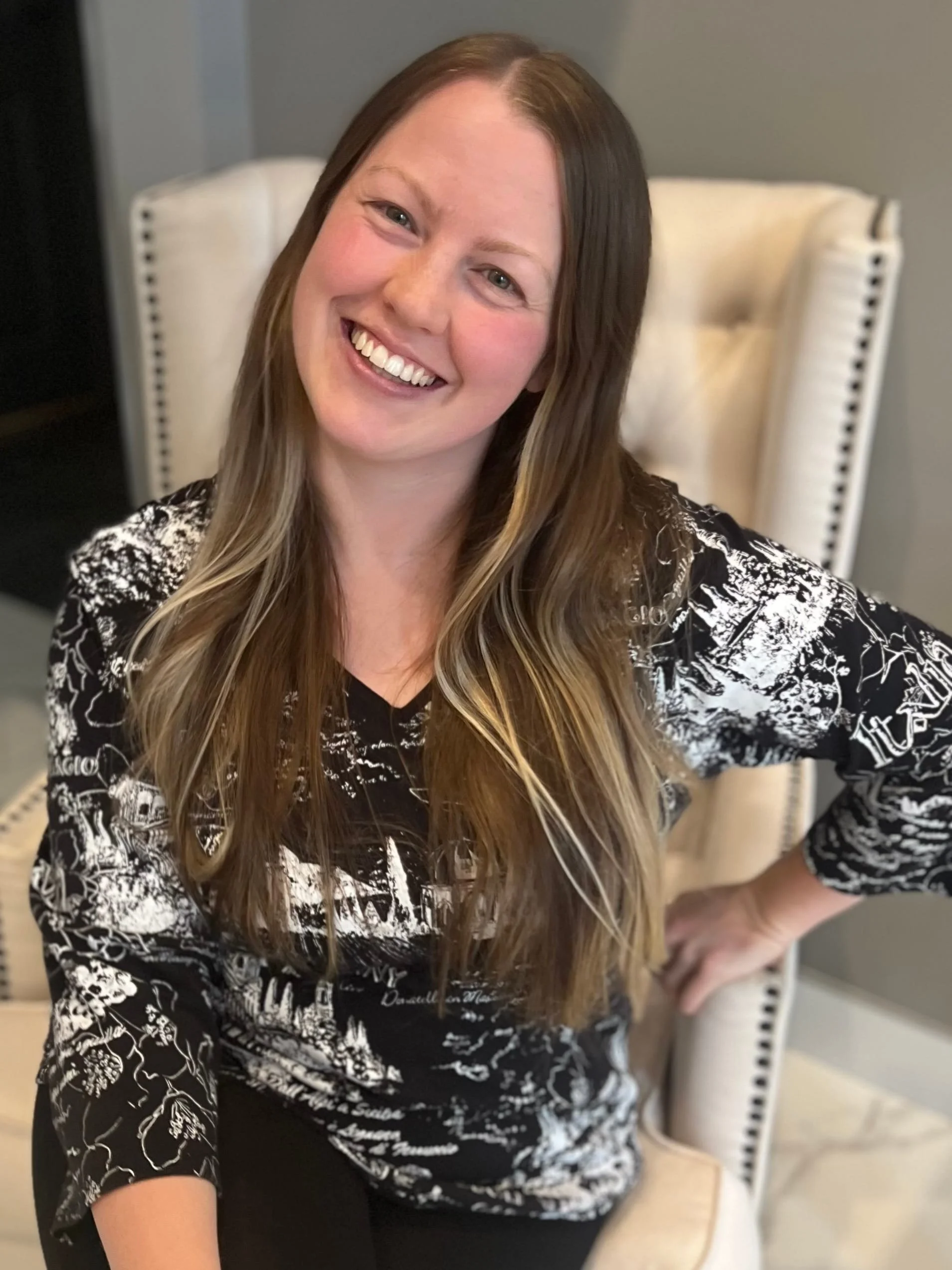 A woman with long light brown hair smiling and sitting on a white chair with nailhead trim, wearing a black and white patterned top.