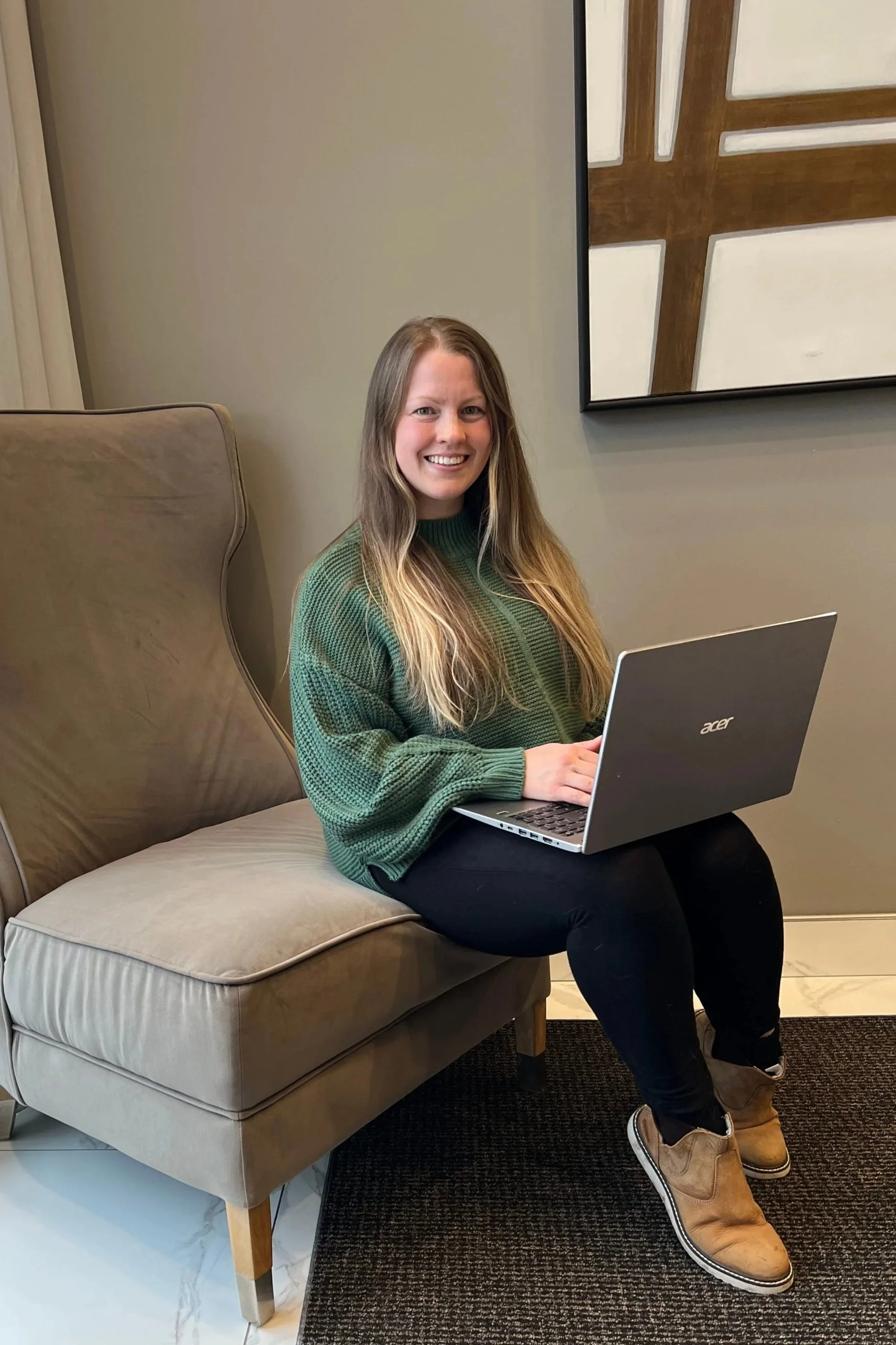 A woman with long hair smiling while sitting on a beige ottoman, using a silver Acer laptop, wearing a green sweater, black pants, and brown boots, in a room with a beige wall, a large framed geometric artwork, and a dark carpeted floor.