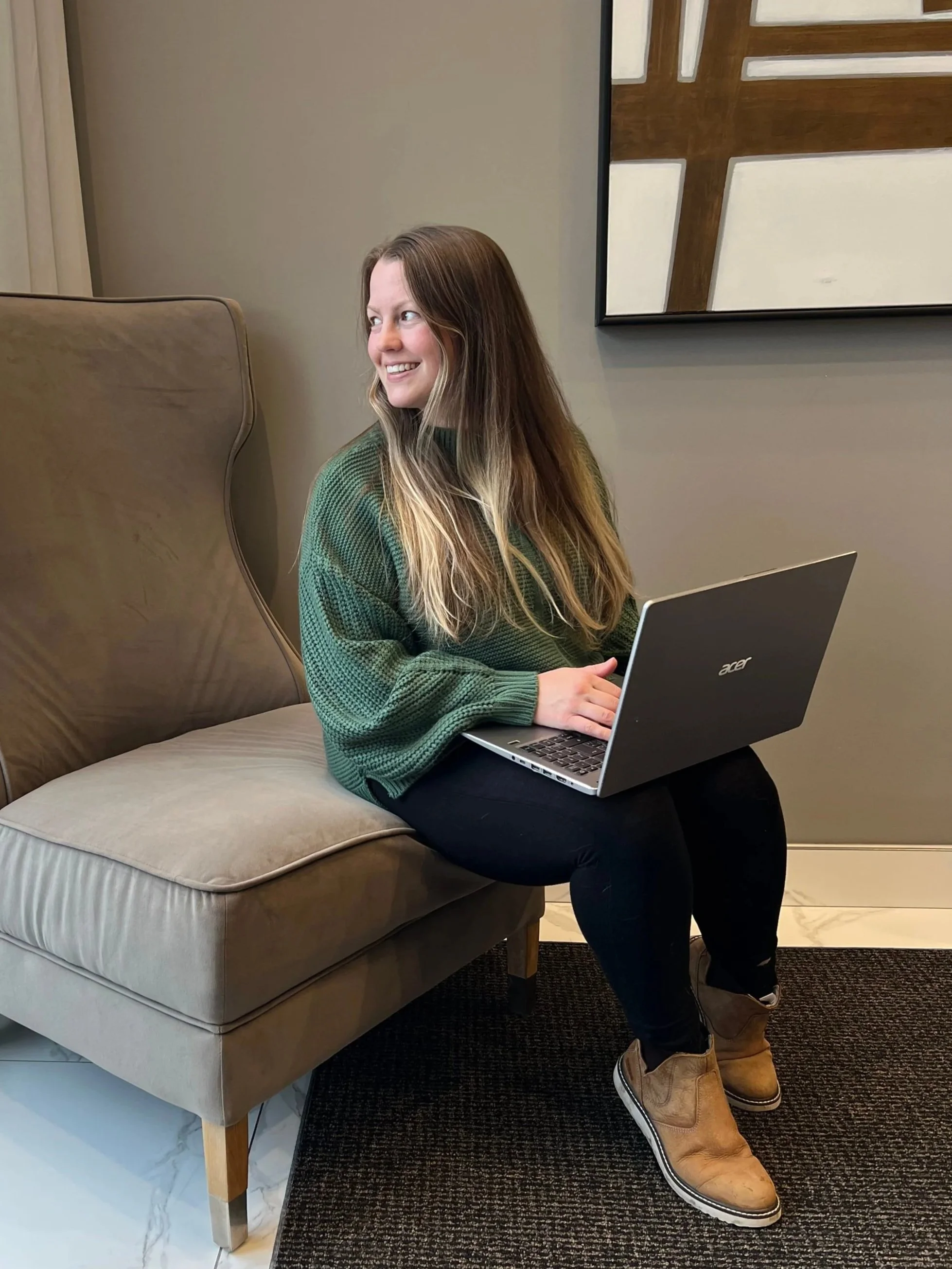 A woman sitting on a beige couch, working on a laptop with a smile, in a room with a wall art and a black rug.