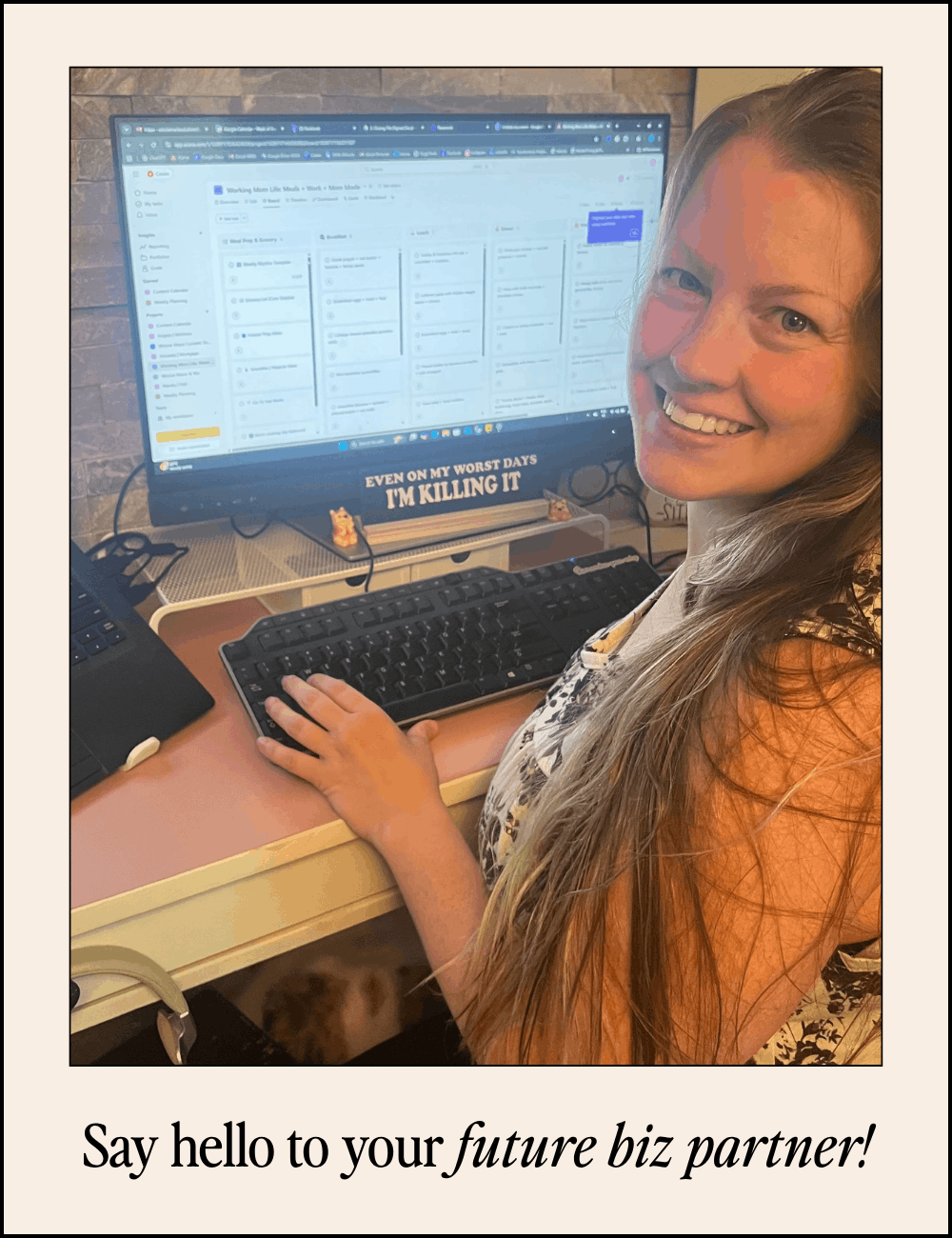 A woman smiling at her computer at a desk, with a large monitor displaying a digital planning or project management tool. There is a keyboard and mouse in front of her, and a motivational sticker on the monitor says, 'Even on my worst days I’m killing it.' The caption reads, 'Say hello to your future biz partner!'