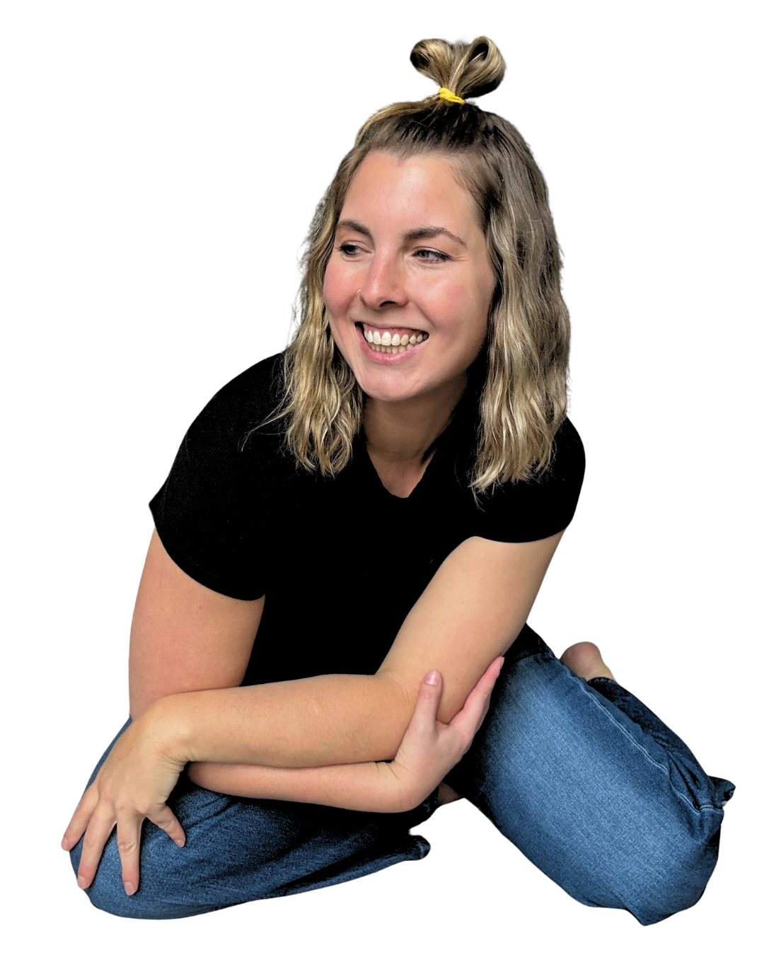 Young woman with wavy blonde hair tied in a top ponytail, smiling, wearing a black t-shirt and blue jeans, sitting on the floor with legs crossed and arms resting on her knees, against a black background.