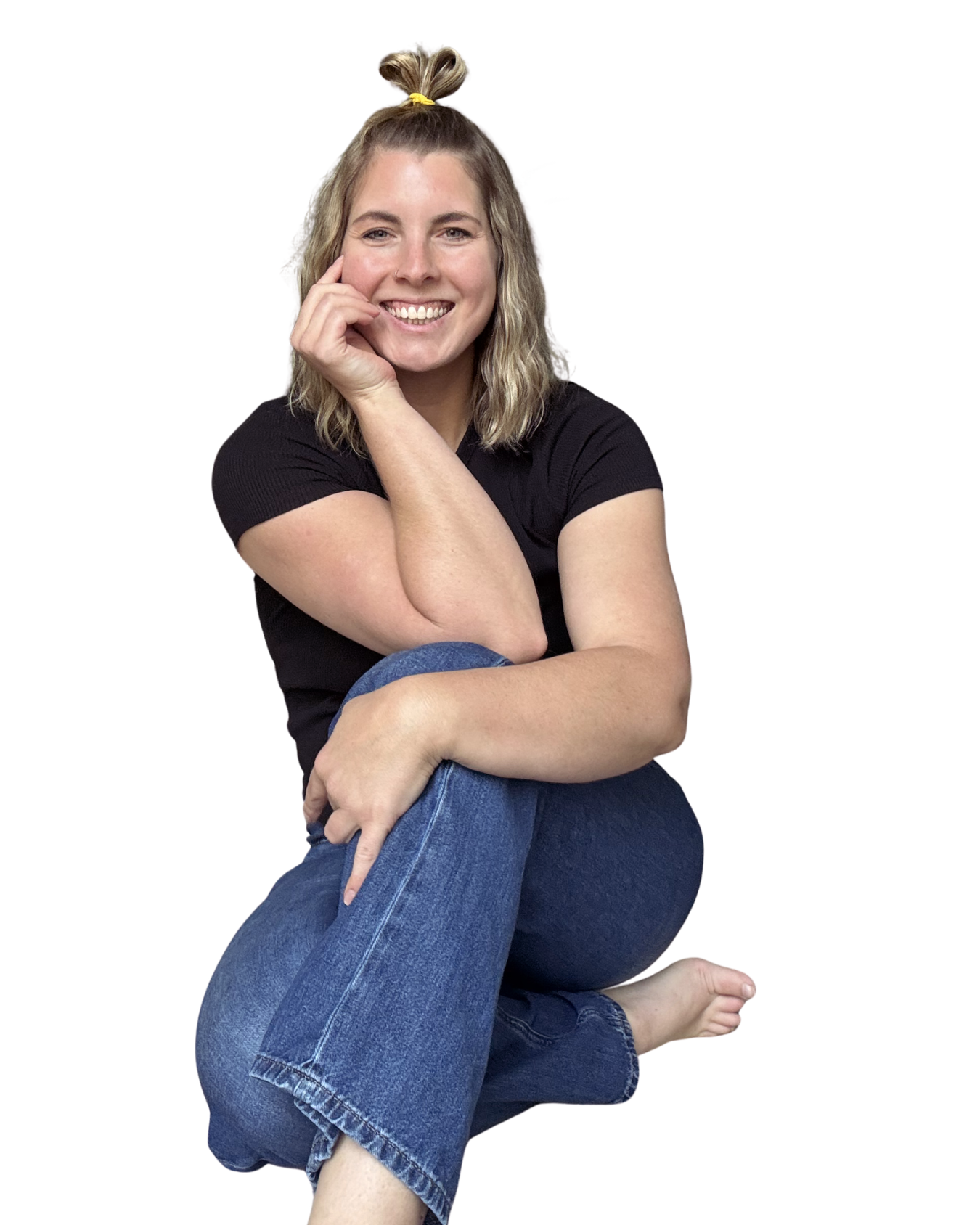 A woman sitting on her knees, smiling, with blonde hair tied up in a bun, wearing a black t-shirt and blue jeans.