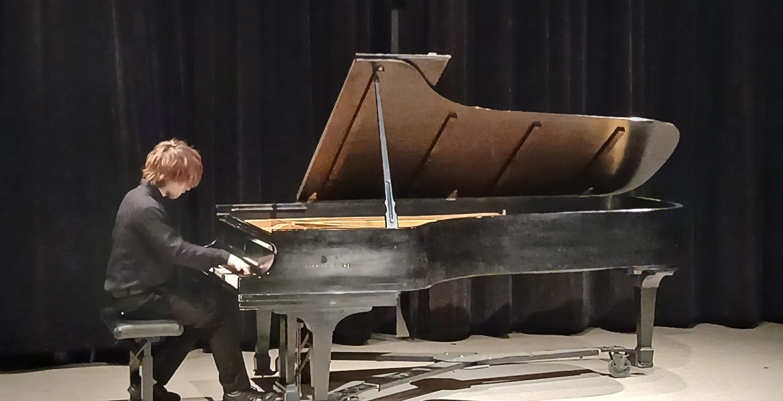 A person with brown hair playing a black grand piano on a stage with black curtains.