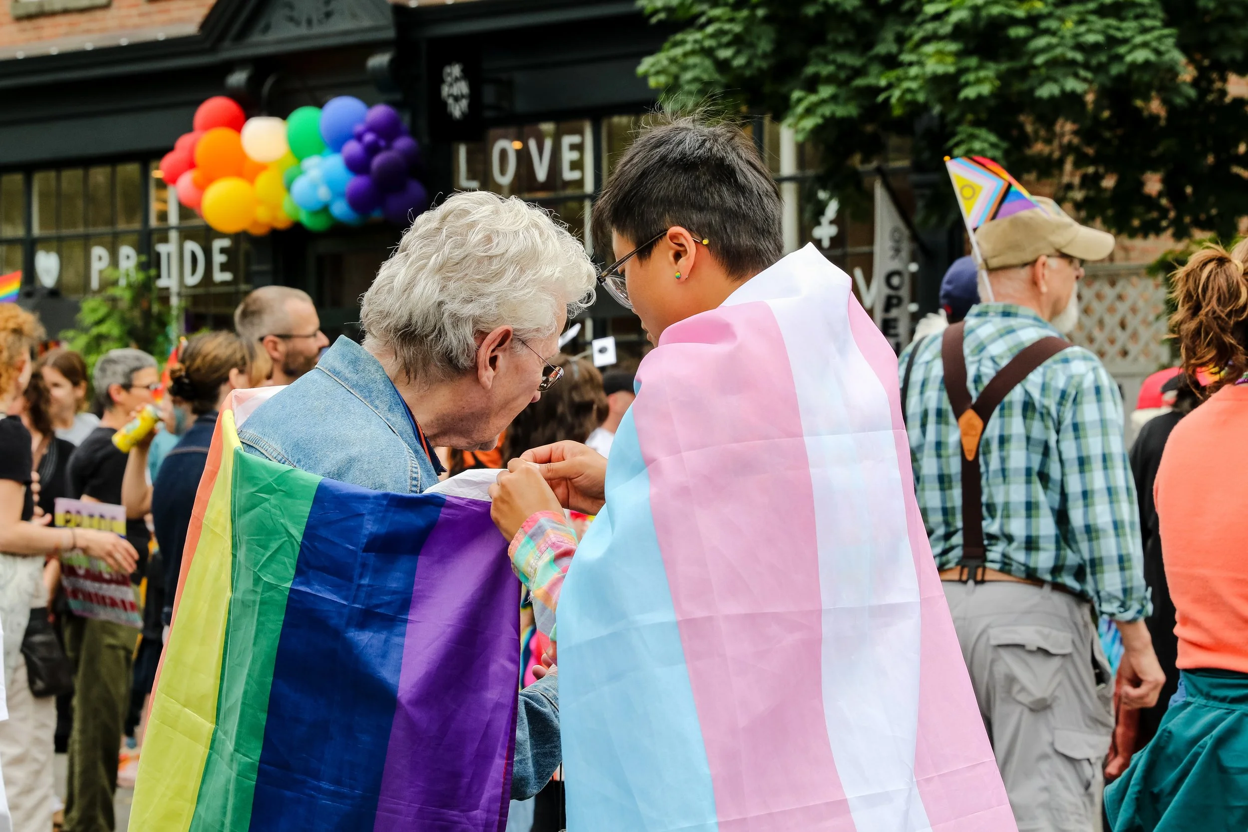 Two people at a pride parade, one older with gray hair and glasses, wrapped in a rainbow pride flag, and one younger with short dark hair and glasses, wrapped in a pastel pink, blue, and white transgender pride flag. They are engaged in a conversation amidst a crowd, with rainbow decorations and signs that say "LOVE" and "PRIDE" in the background.