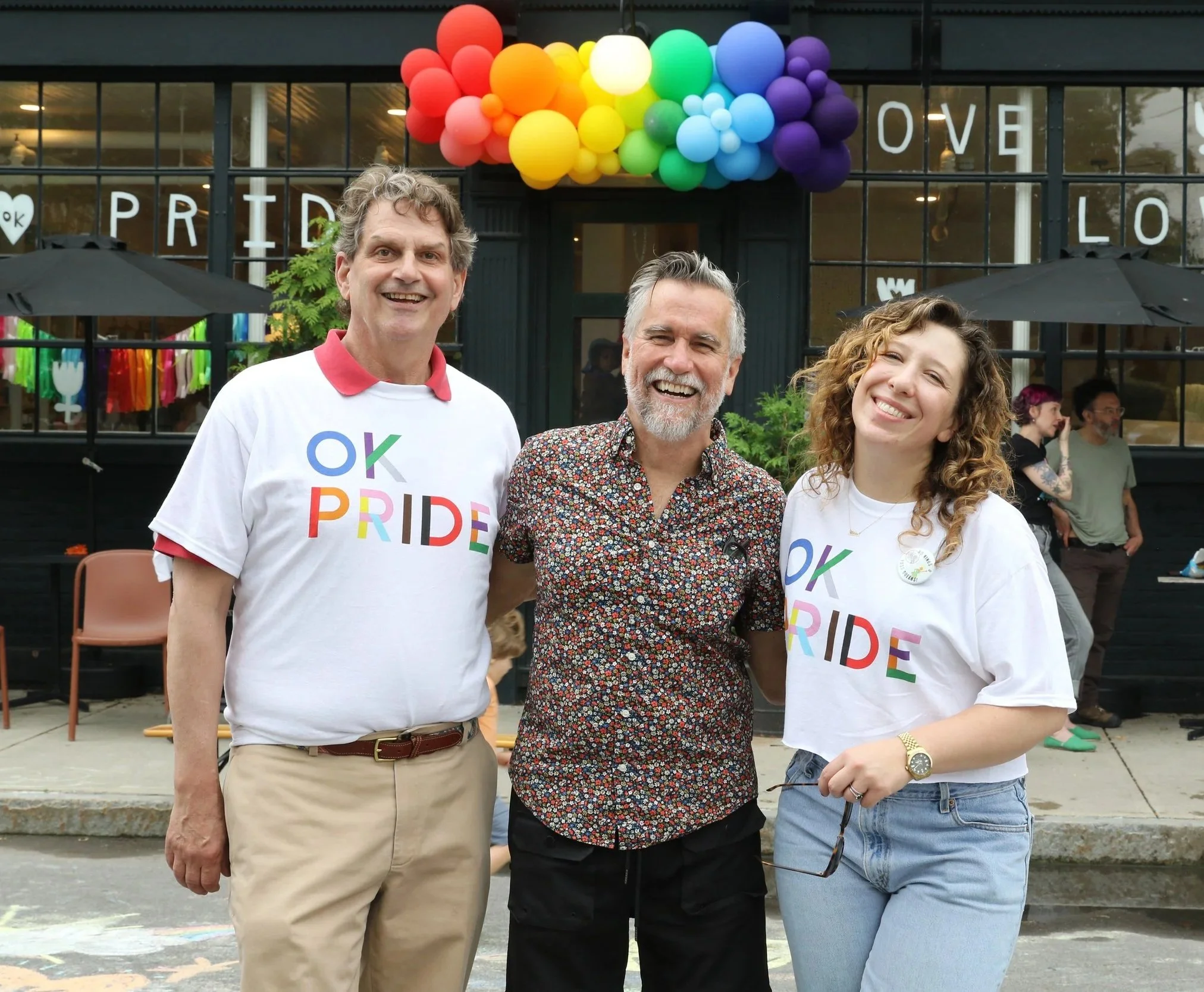Three people smiling outdoors during a Pride event, two wearing white T-shirts with rainbow "OK PRIDE" text, standing in front of a building decorated with rainbow balloons and Pride signage.